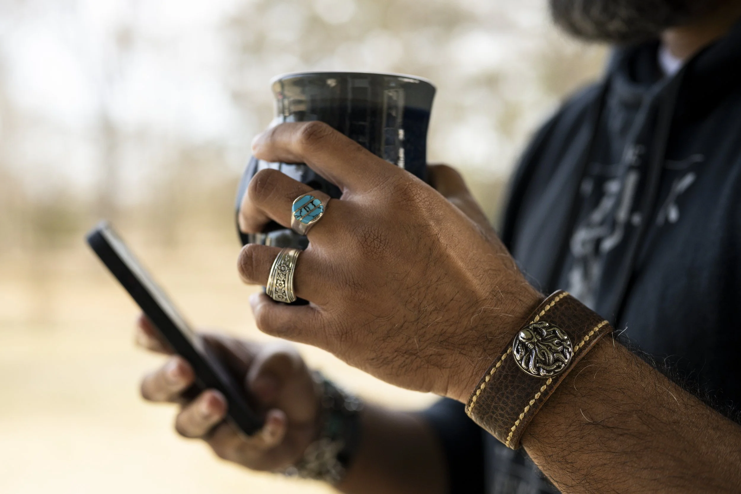 Close-up of a person holding a smartphone with both hands, wearing multiple rings and a leather bracelet, with a blurred background.