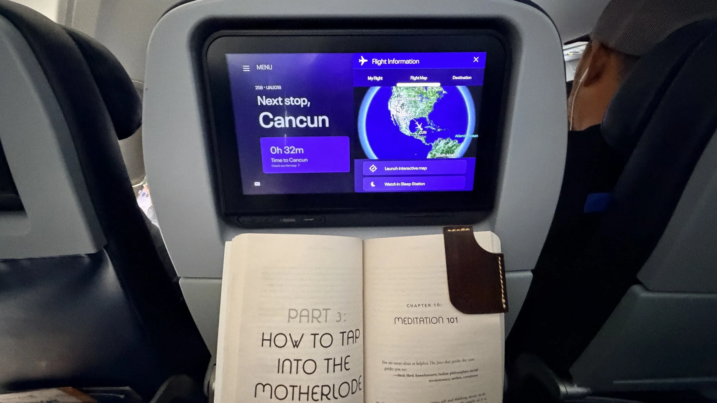 An airplane seatback screen shows a flight approaching Cancún, Mexico, with 32 minutes until arrival and the route displayed on a map. A book rests on the tray table below.