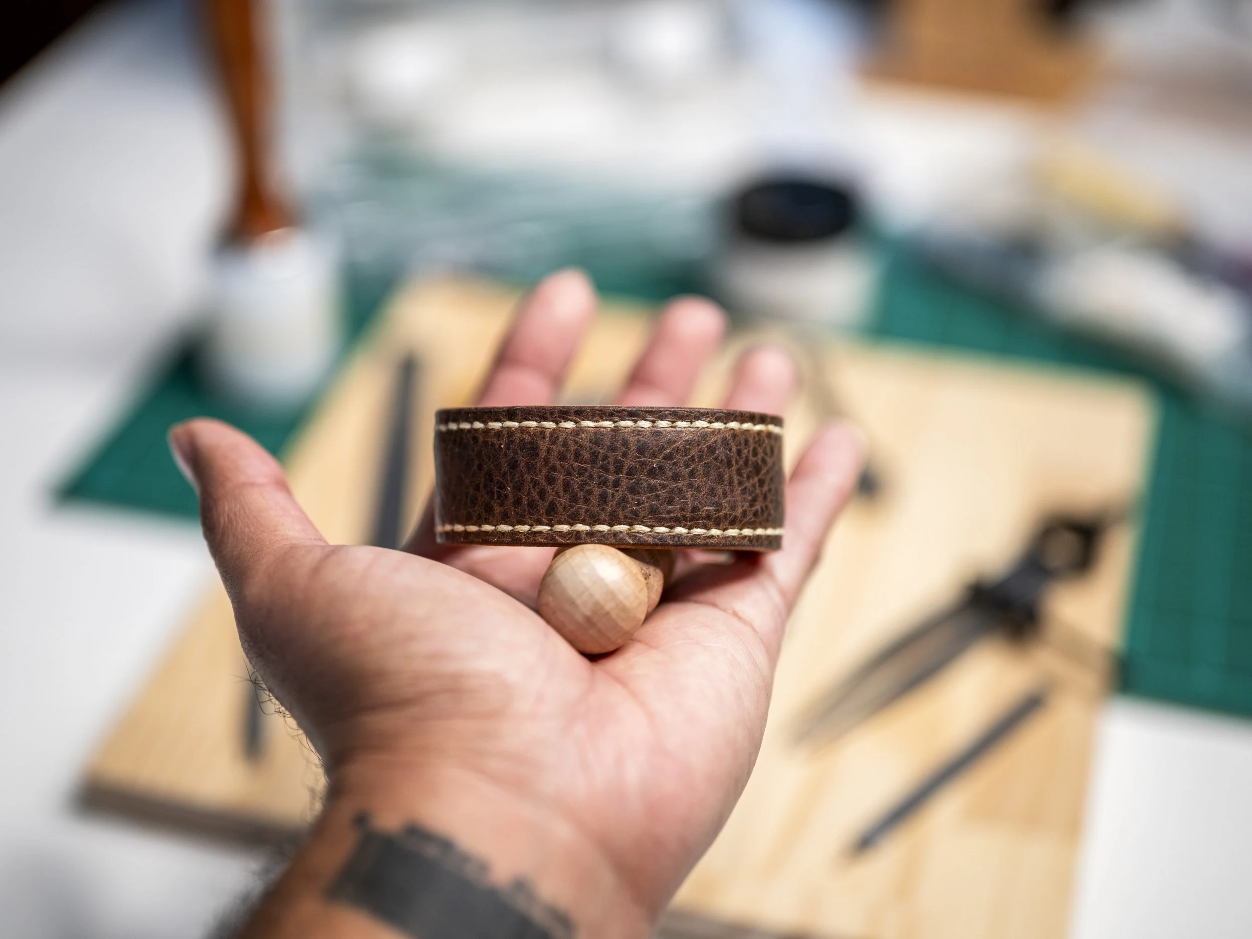 A Black hand holds a handcrafted brown leather cuff to show its finished back, with neat contrasting stitching around the edges and the workshop softly blurred behind it.
