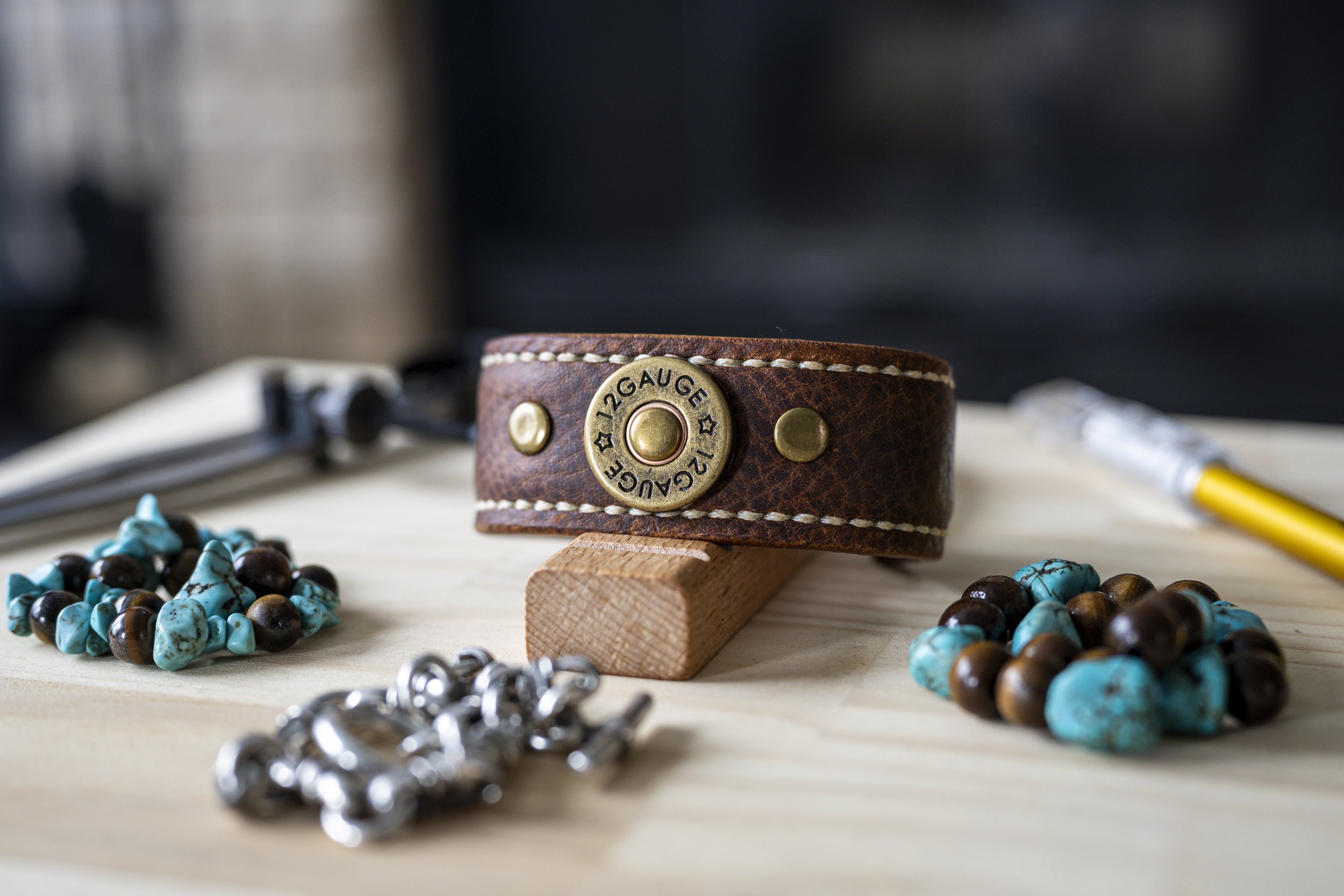 The Koa handcrafted brown leather cuff with its round brass-toned concho and contrasting stitching is displayed on a wooden stand, surrounded by turquoise-colored beads and silver-toned materials on the workshop table.