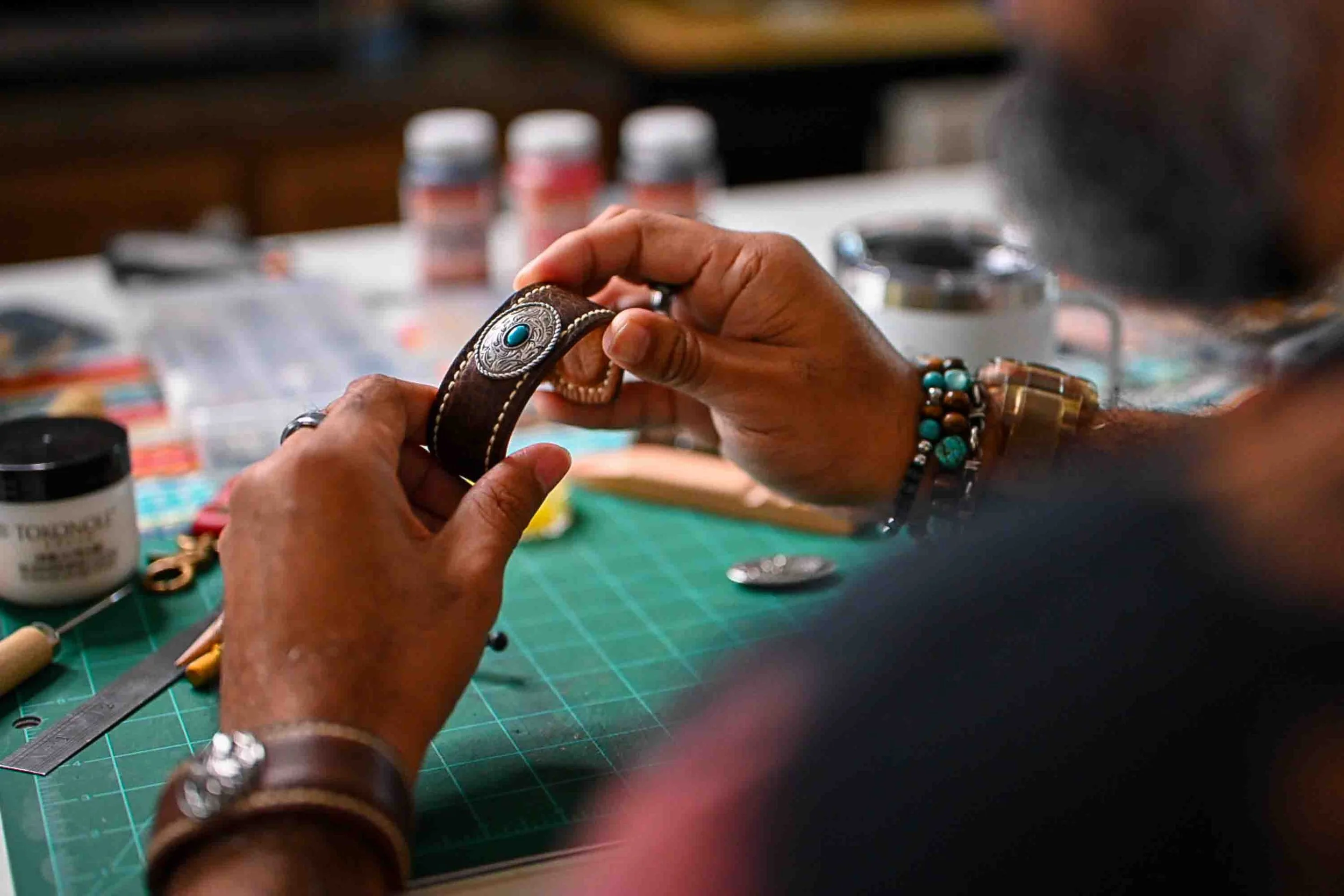 Hands hold the Moana leather cuff over a green cutting mat at a leatherworking bench, surrounded by tools and craft supplies while the piece is being worked on.
