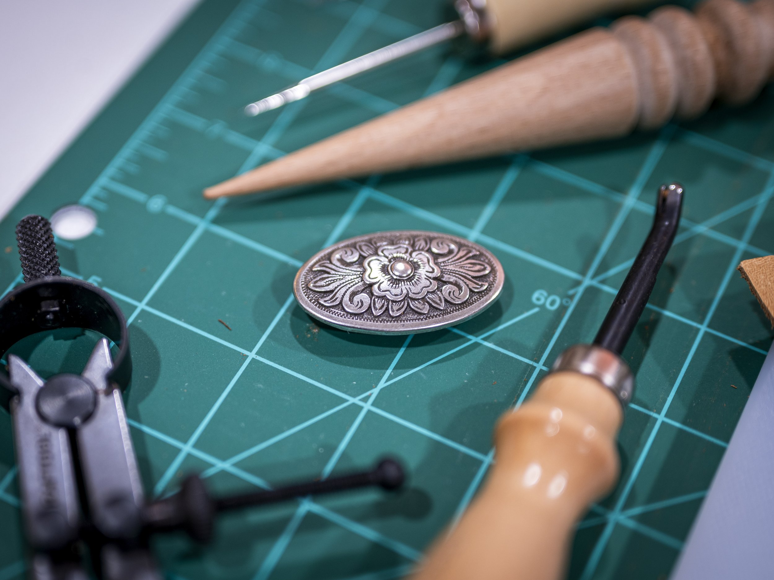 Close-up of crafting tools on a green cutting mat, including a decorative metal button, a craft knife, a sewing pin, and a partially visible leather or fabric item.