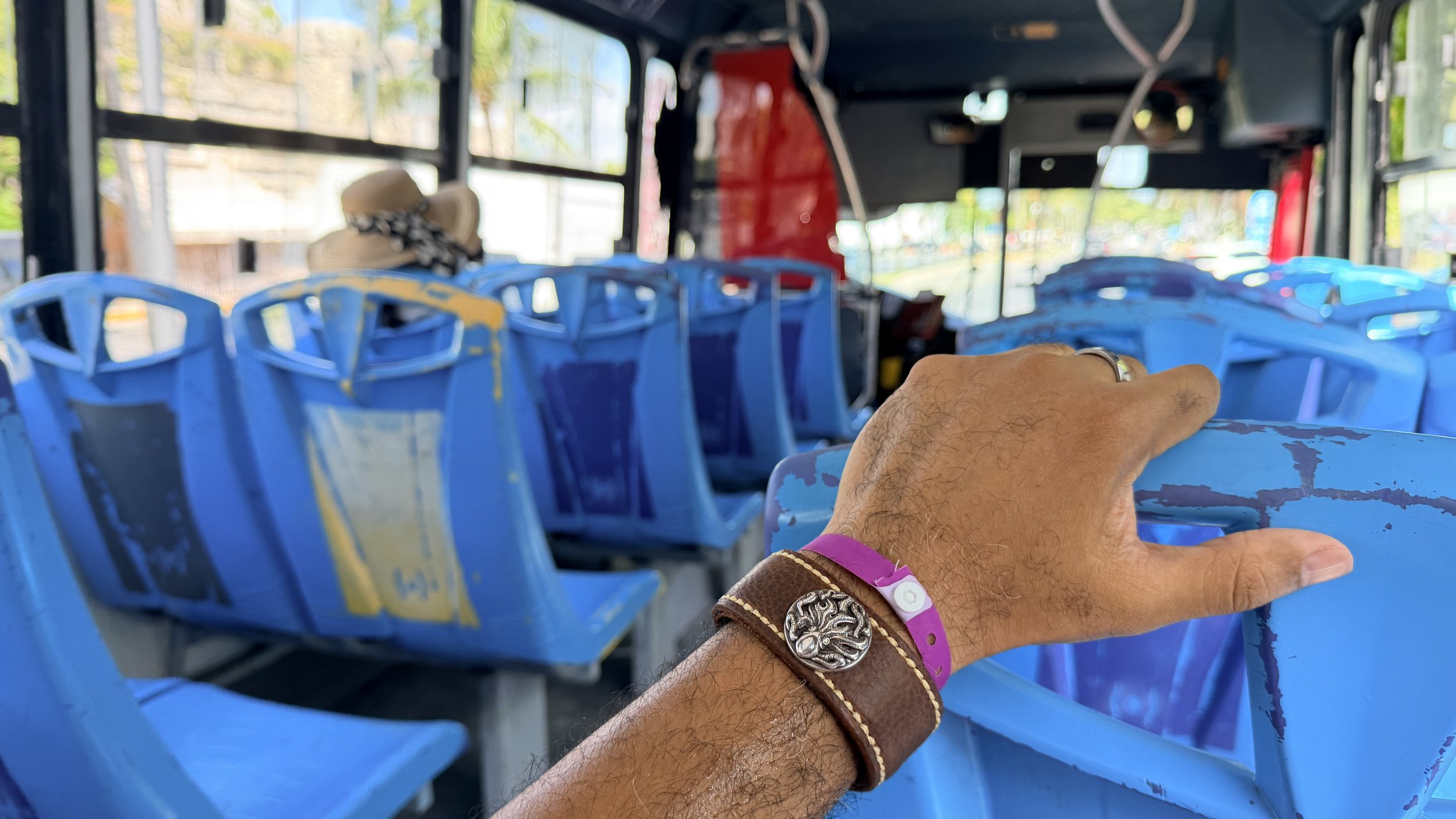 A Black man’s hand wearing the Hilo handcrafted brown leather cuff rests across a bright blue seat while riding a bus, with rows of blue seats extending toward the front.