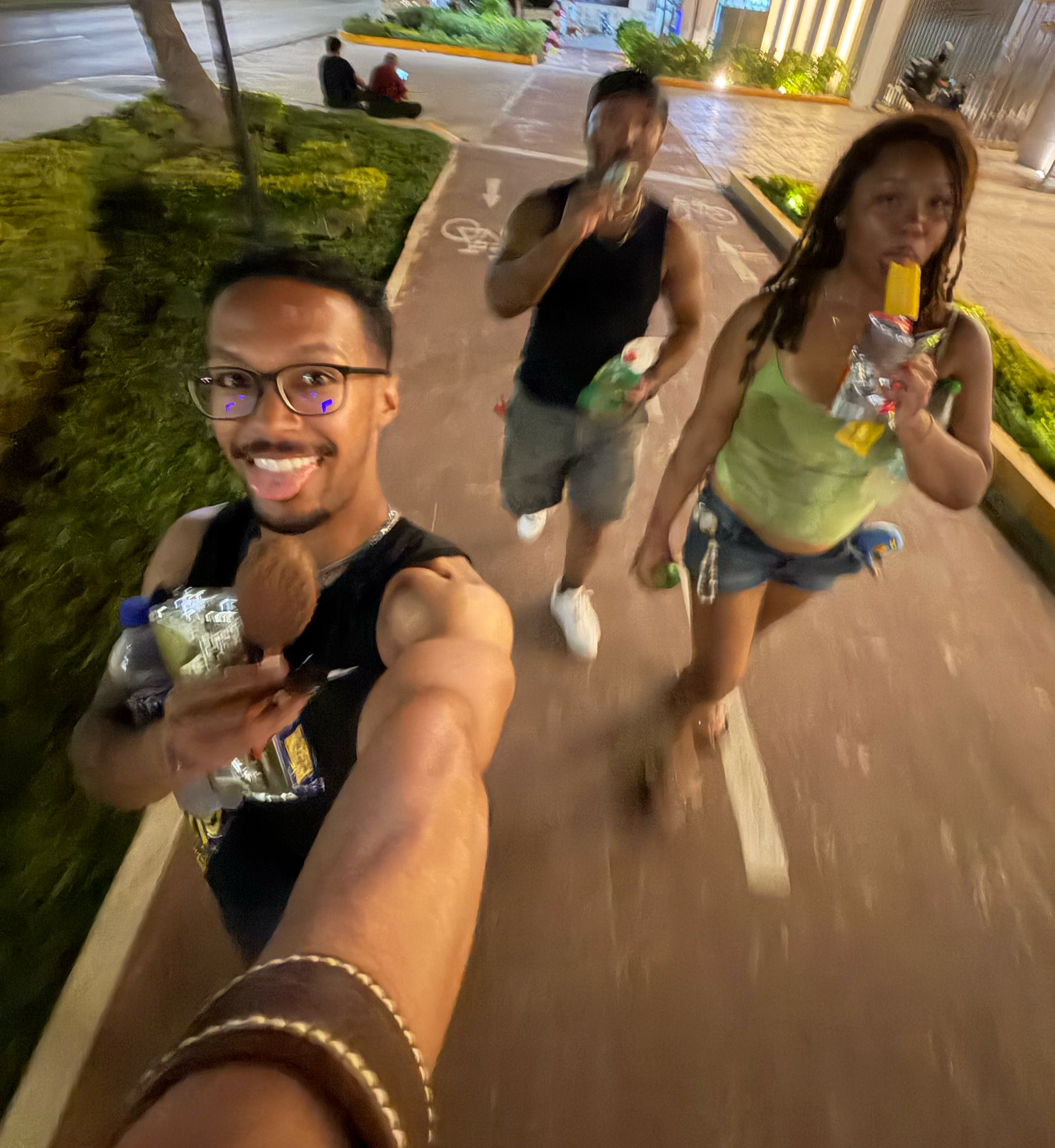A smiling Black man wearing the Makahā leather cuff takes a wide-angle selfie with two friends of color during an evening out in Cancún, Mexico, along a brightly lit outdoor walkway surrounded by tropical greenery.