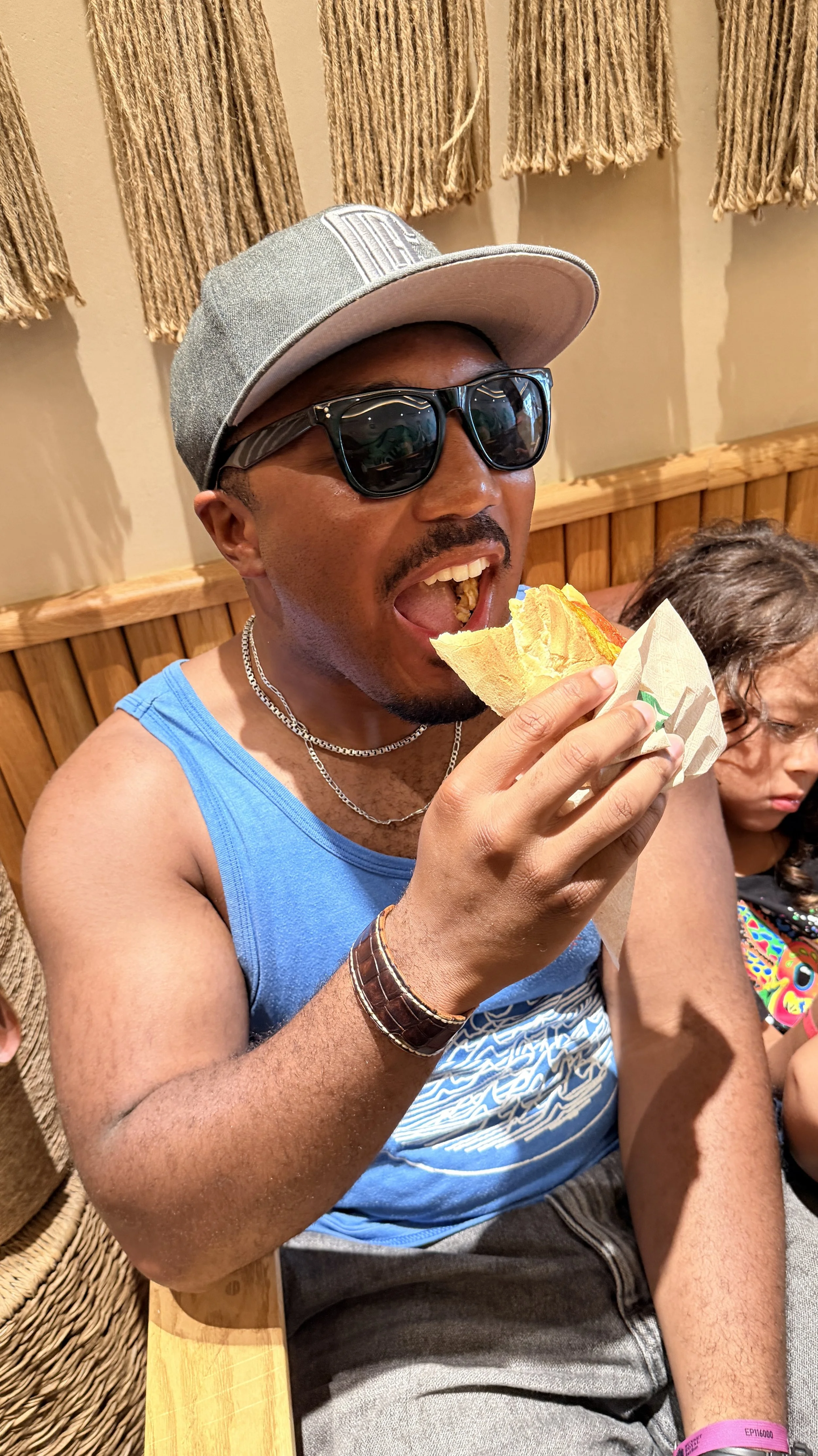 A Black man wearing sunglasses, a gray cap, blue tank top, and the Makahā handcrafted leather cuff smiles as he prepares to take a bite of a taco at an outdoor restaurant.