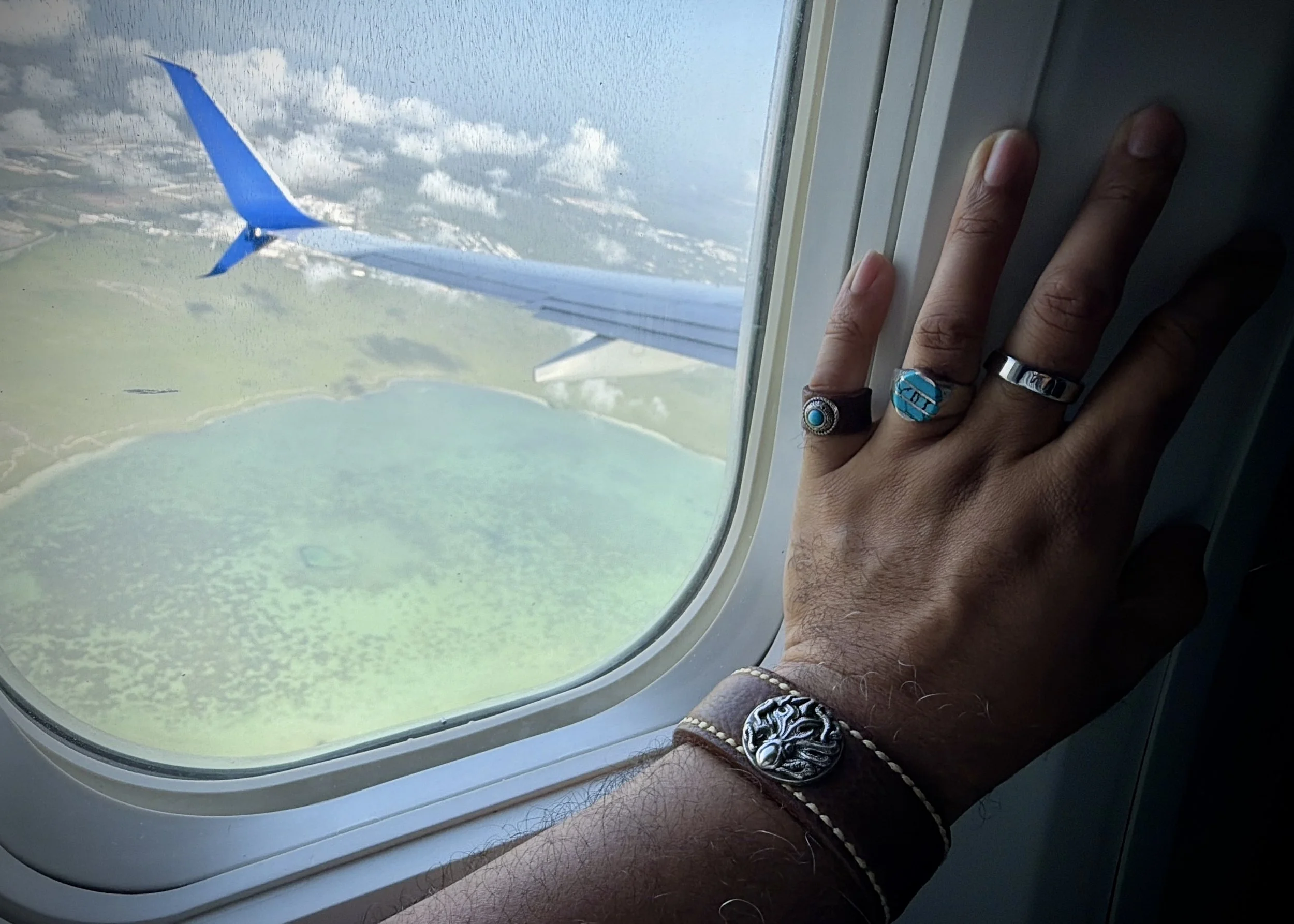 A hand wearing the Hilo leather cuff and turquoise rings rests beside an airplane window, with the wing, blue sky, clouds, and turquoise Caribbean water visible below on the flight to Cancún, Mexico.