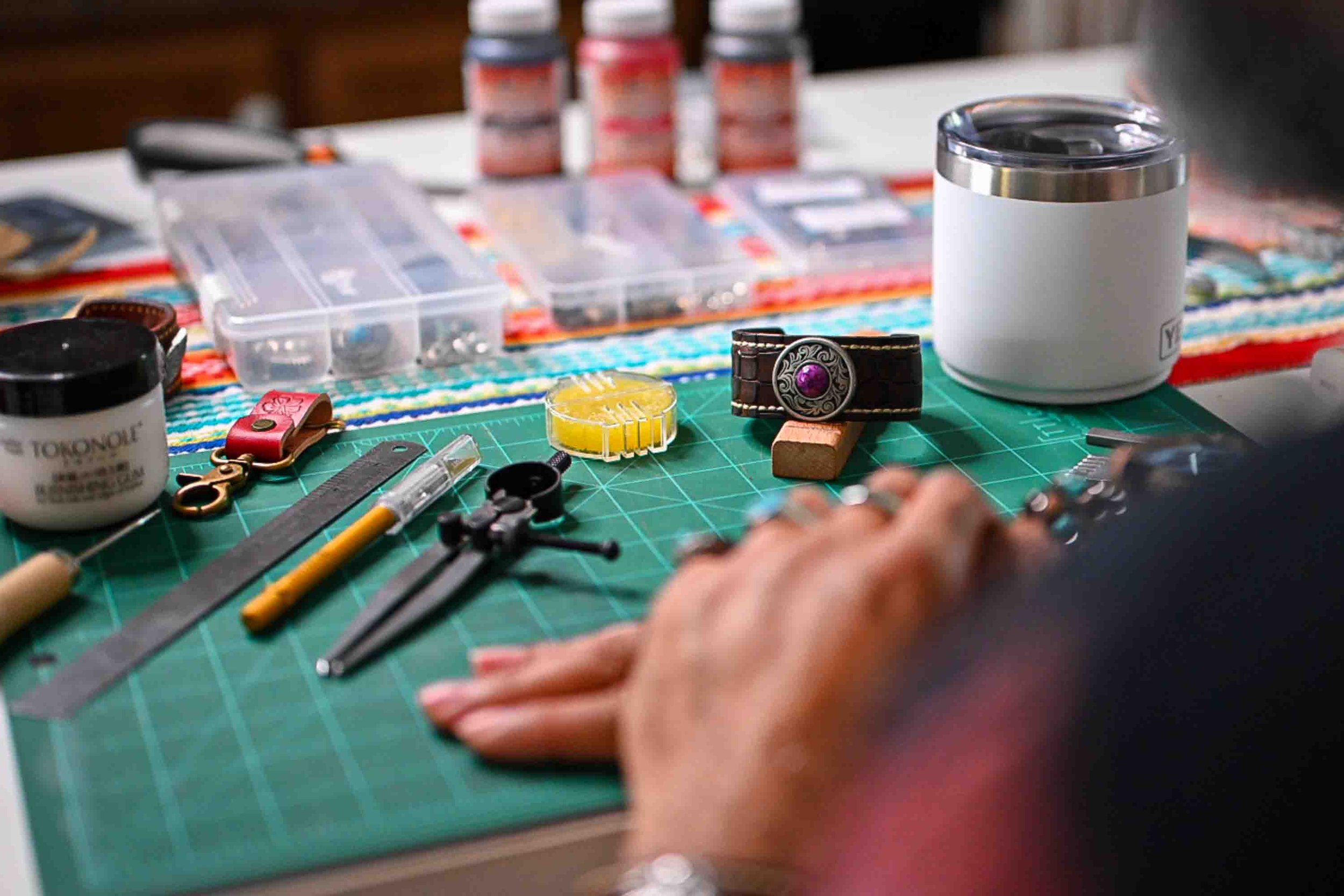 A close-up of various crafts supplies on a green cutting mat, including jewelry pieces, a measuring ruler, and a person's hand working on a jewelry project.