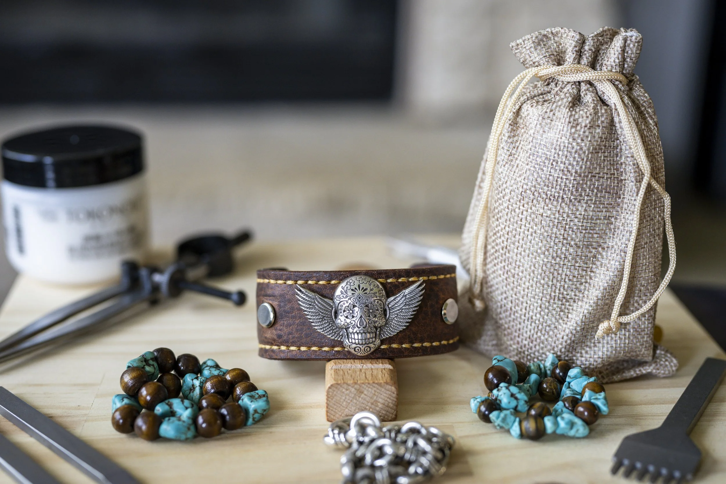 A collection of jewelry and accessories on a wooden surface, including turquoise and brown beaded necklaces, a leather wristband with a skull and wings design, a silver ring, a small burlap drawstring pouch, and blurred objects in the background.
