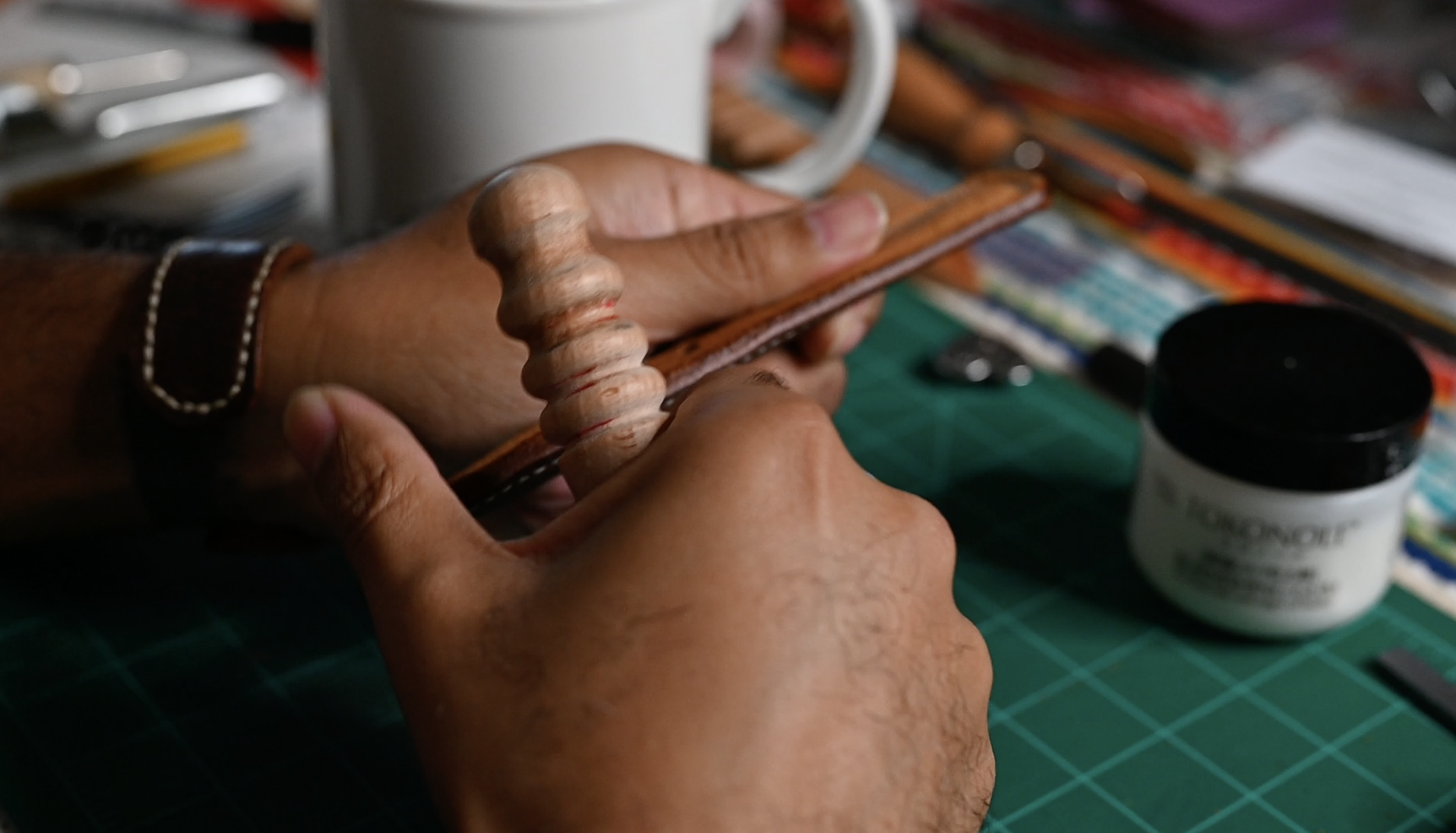 Hands burnish the edge of a small brown leather piece at a leatherworking bench, with a green cutting mat, hand tools, adhesive, and a coffee mug among the workshop supplies.