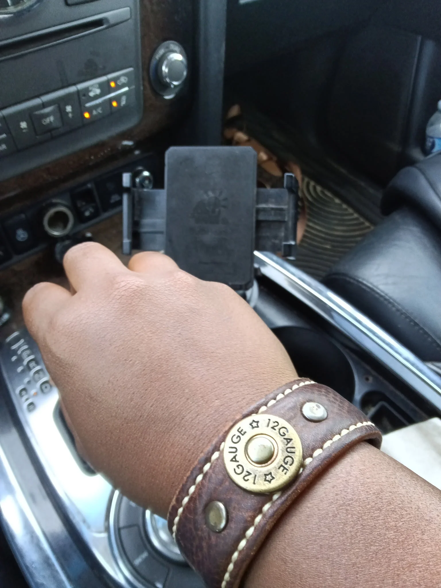 A Black woman’s hand wearing the Koa handcrafted brown leather cuff adjusts a smartphone mounted beside the dashboard inside a vehicle.