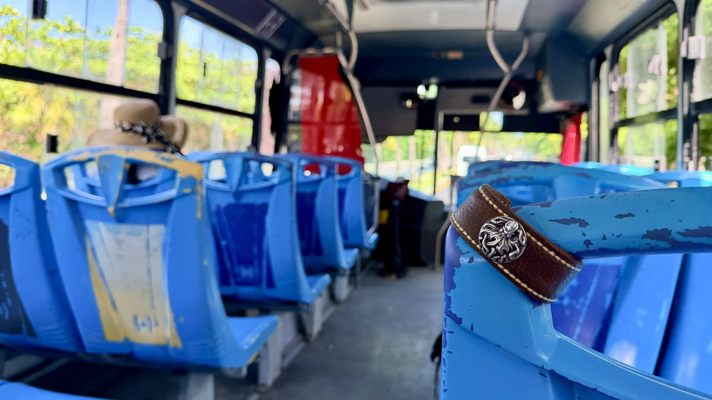 The brown leather Hilo cuff with its octopus concho is draped over the back of a bright blue seat inside a local bus, with rows of blue seats, open windows, and lush green trees visible outside during a ride through Cancún, Mexico.