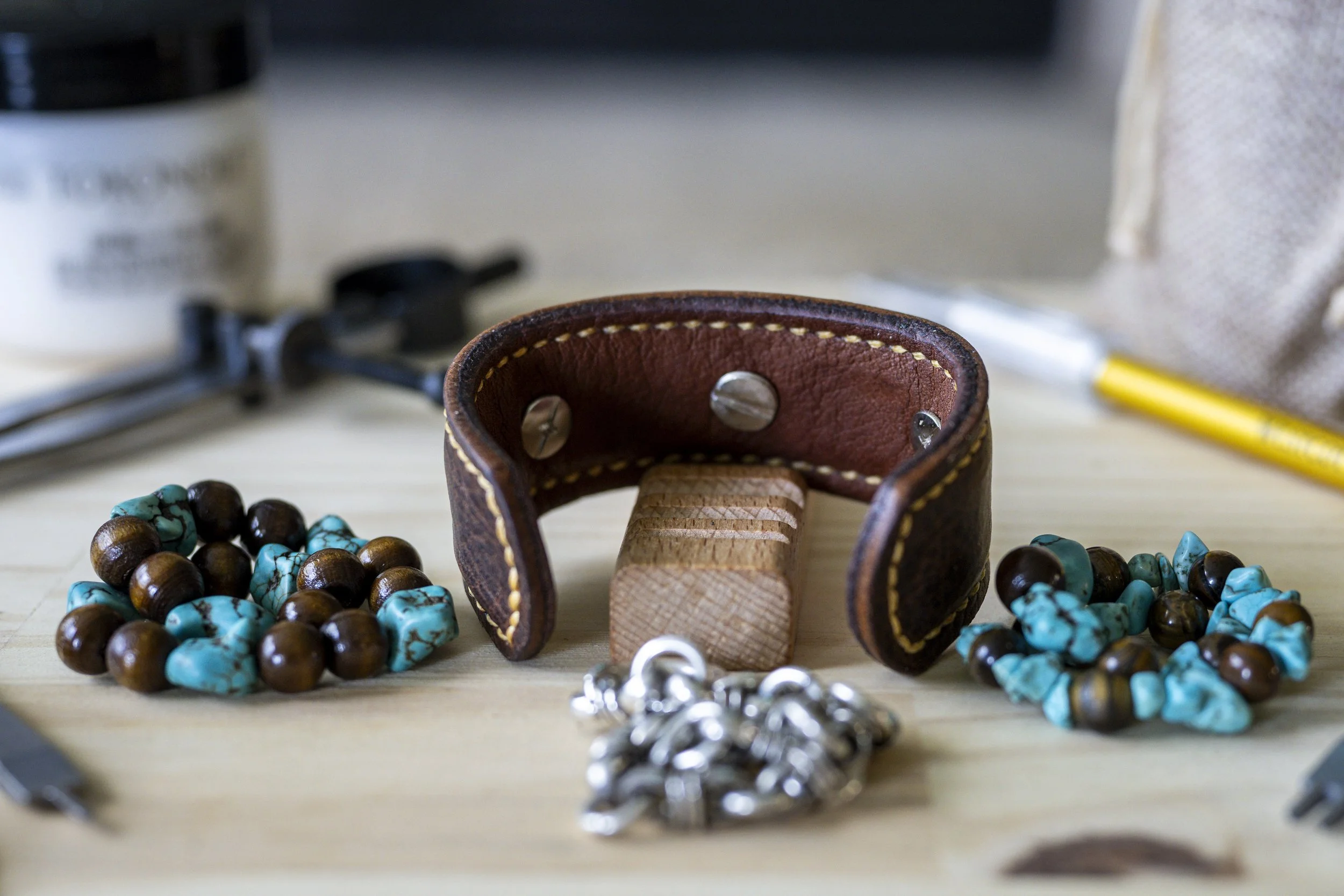 Close-up of a leather bracelet with yellow stitching and metal snaps, surrounded by turquoise and brown beaded necklaces and jewelry on a light wooden surface, with tools and fabric in the background.