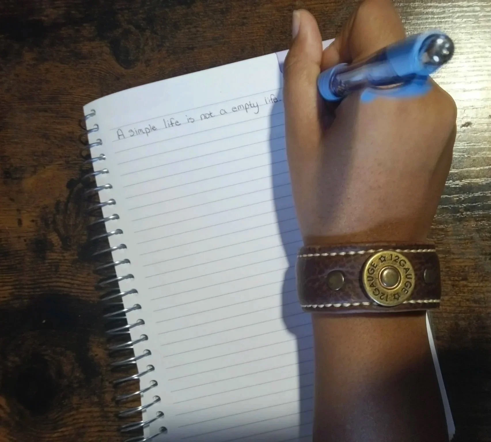 A Black hand wearing the Koa handcrafted brown leather cuff writes “A simple life is not an empty life.” in a spiral notebook with a blue pen at a wooden table.