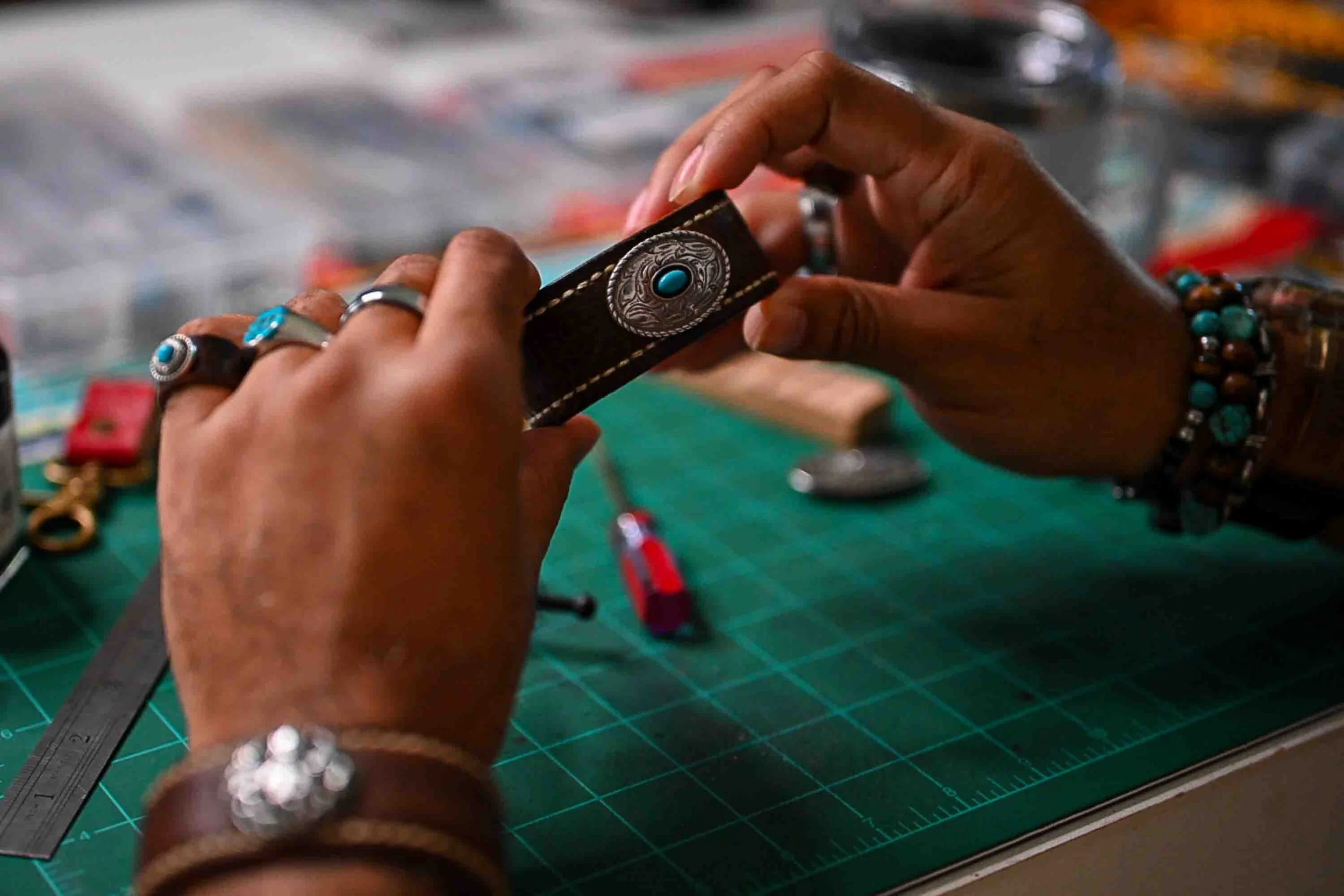 A Black leather artisan works on the handcrafted Moana cuff at a workshop table, adjusting its oval silver-toned concho with turquoise-colored center, surrounded by jewelry and leatherworking supplies on a green cutting mat.