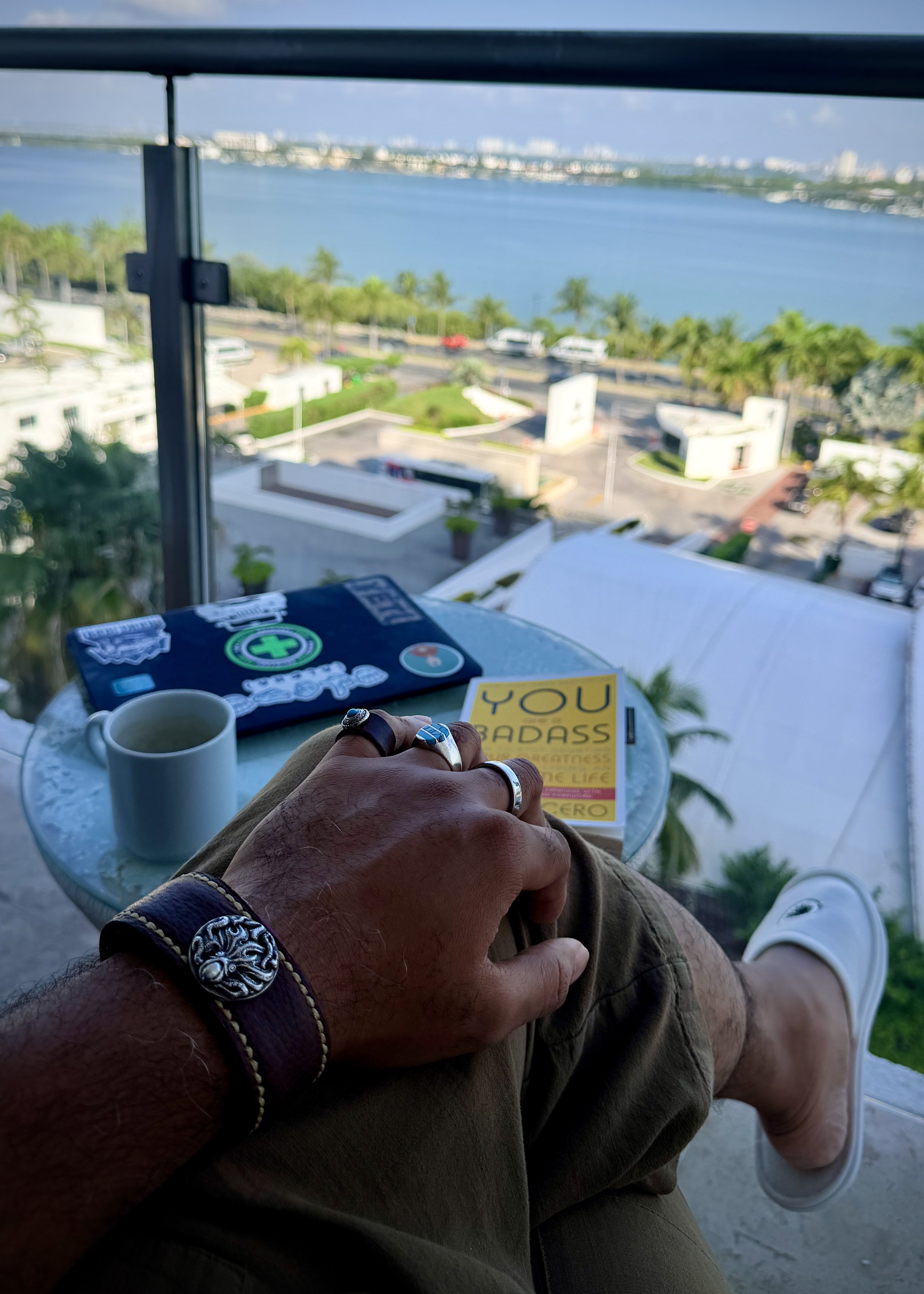 A person sitting at a table with a view of a bay or ocean in the background. The person is wearing bracelets and rings, and has a white mug, a laptop with stickers, and a yellow notebook or journal on the table.