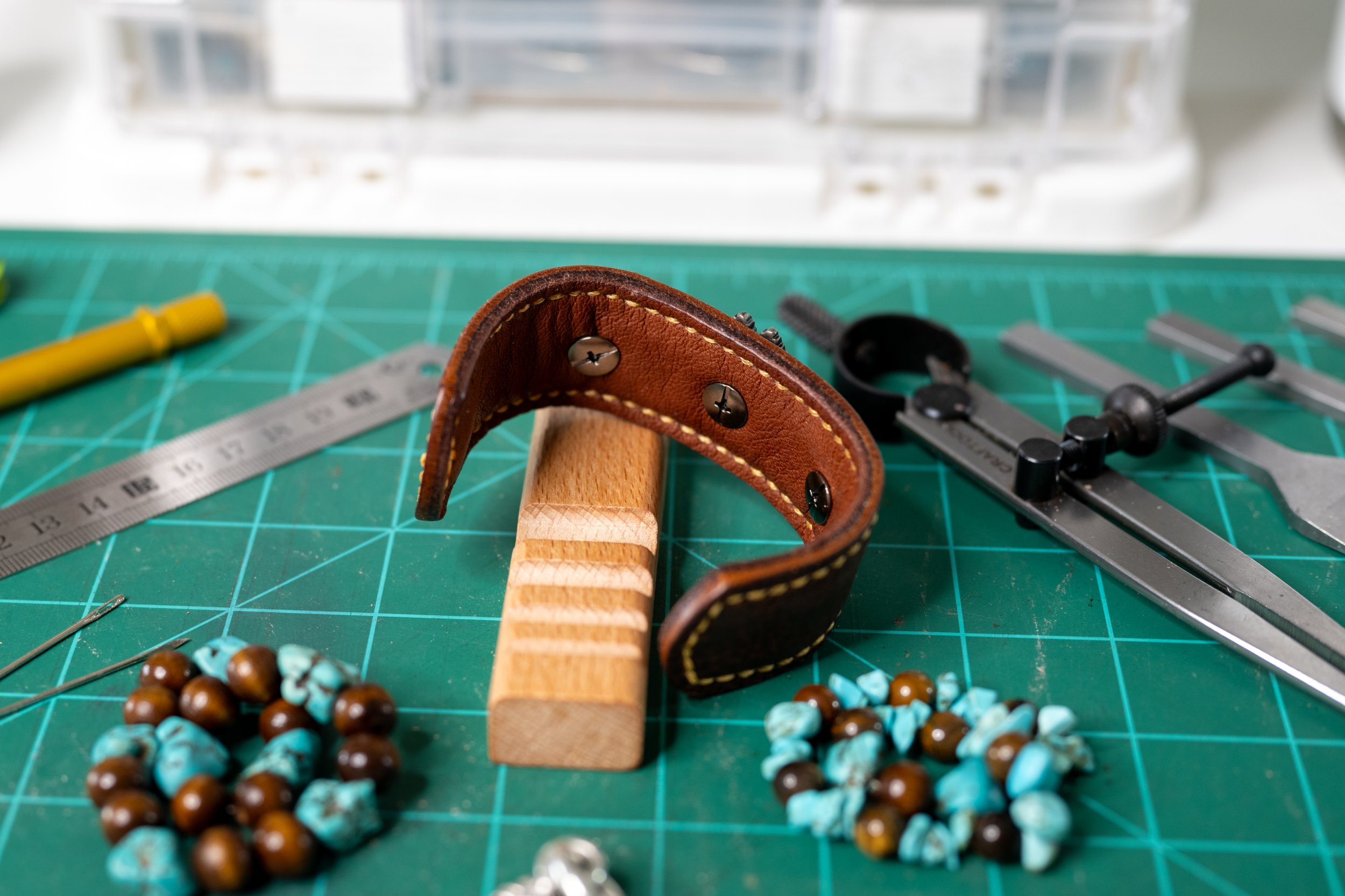 Close-up of crafting tools and accessories on a green cutting mat, including a leather strip, a metal ruler, a compass, beaded jewelry with brown and turquoise beads, and a cutting tool, in a workshop setting.
