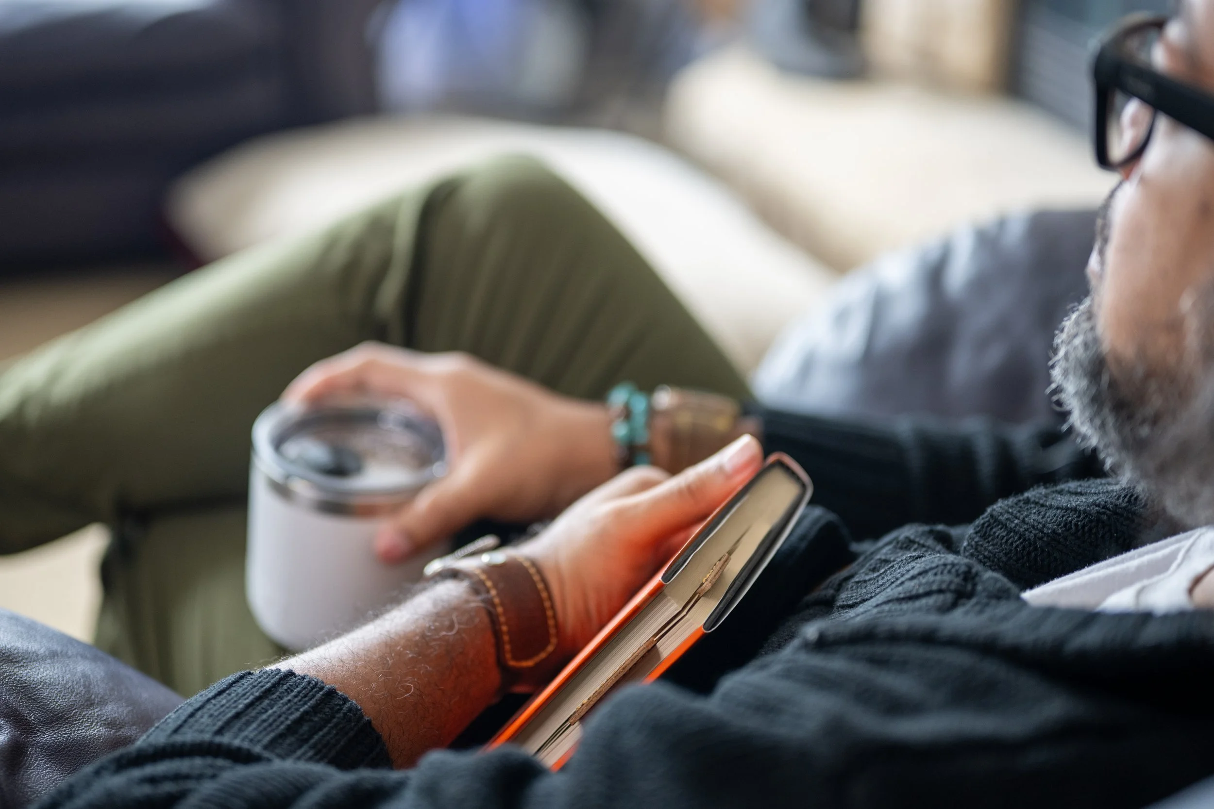 A person with glasses using a smartphone, holding a clear plastic cup with a drink, sitting with crossed legs, wearing dark clothing and a bracelet, with a coffee mug nearby.