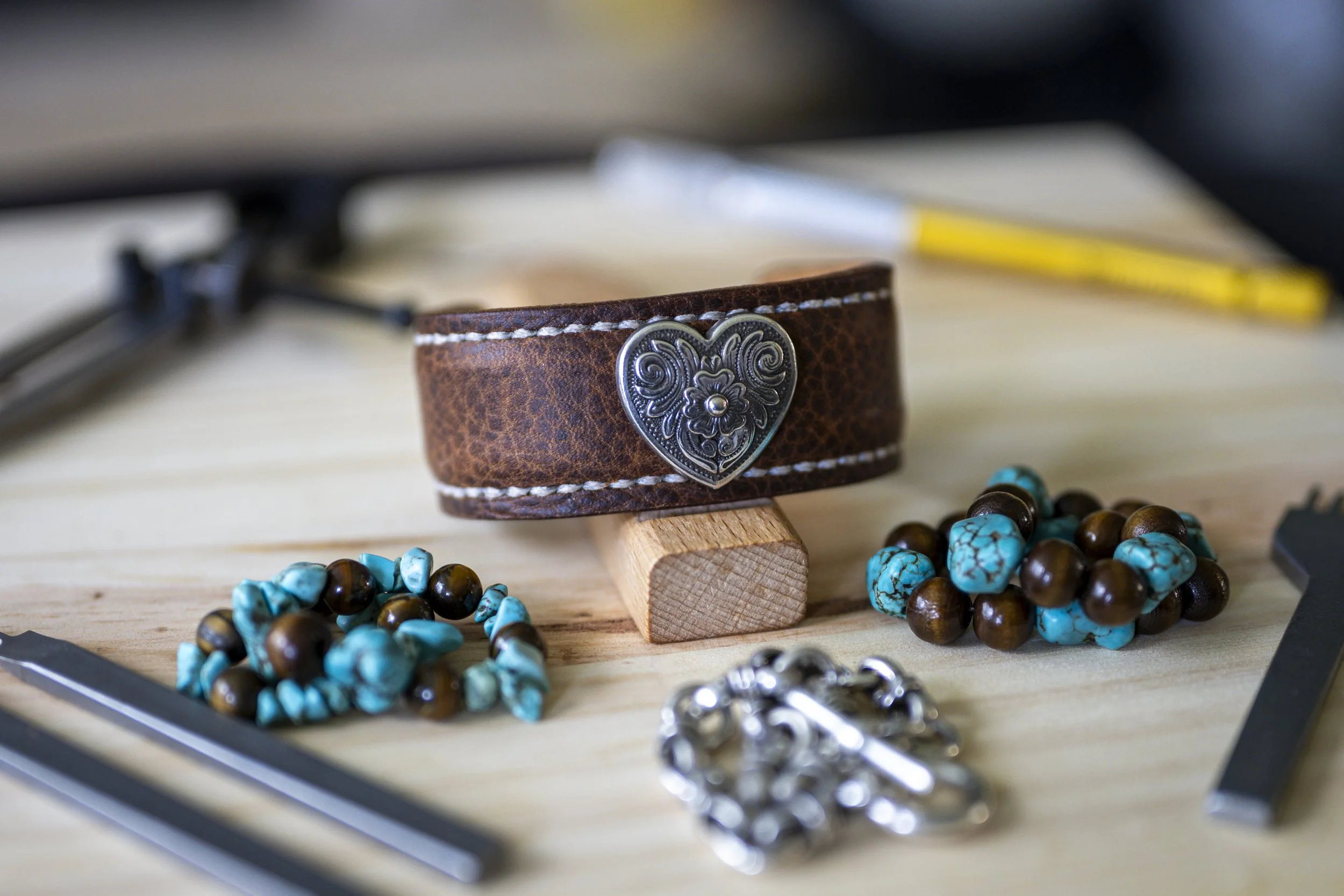 The handcrafted Waimea brown leather cuff displayed on a small wooden stand, featuring white stitching and an ornate silver-toned heart concho, surrounded by turquoise, brown, and silver-toned jewelry on a light wooden workbench.