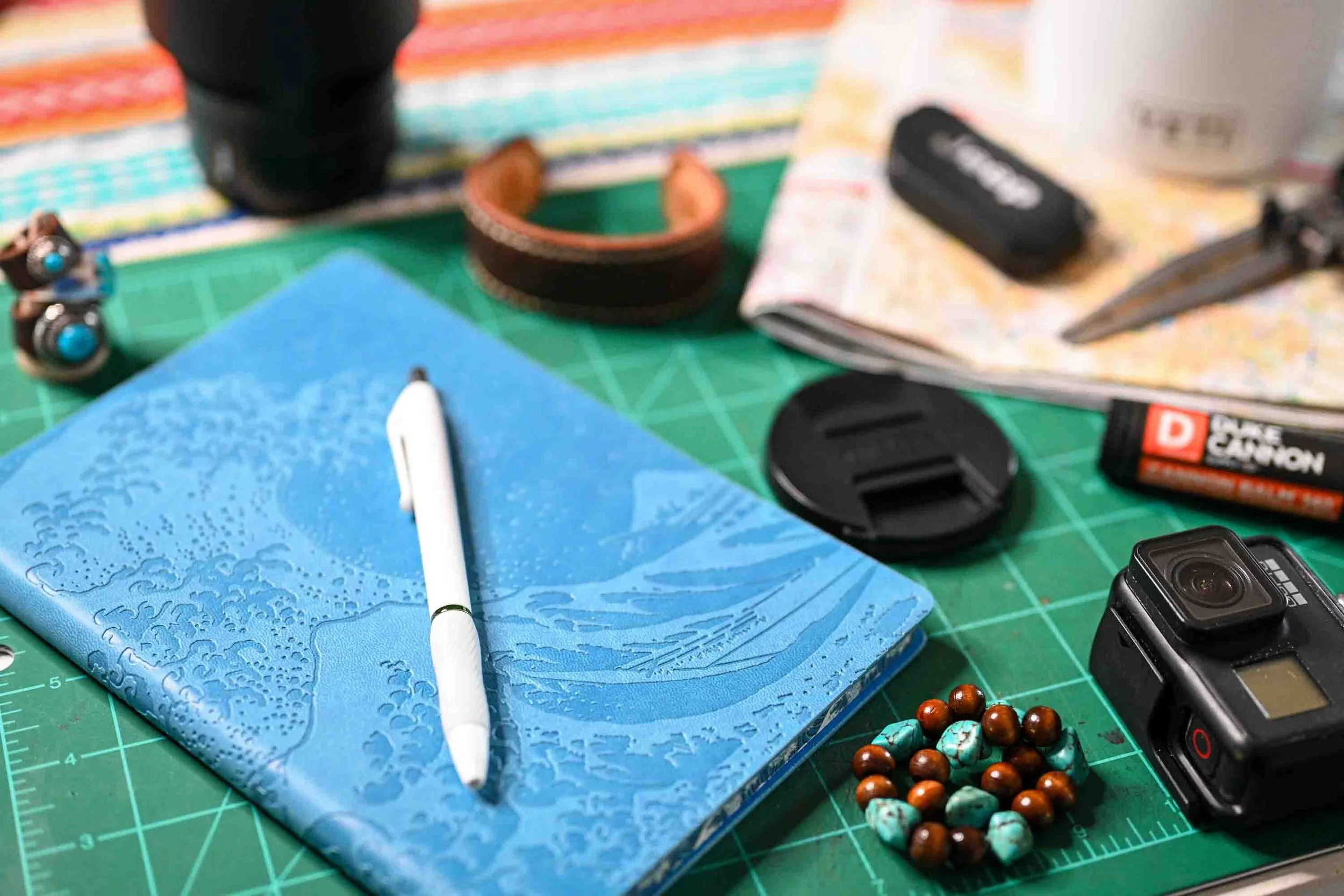 A busy creative worktable with a blue notebook and white pen surrounded by leatherworking supplies, camera gear, electronics, a beaded bracelet, and a coffee mug on a green cutting mat.