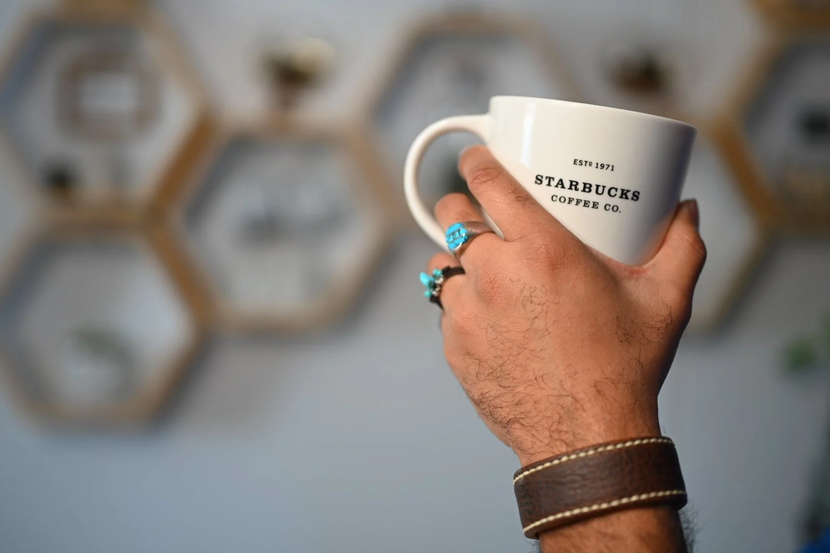 A hand wearing the Makahā leather cuff and turquoise rings holds a white coffee mug in a café, with geometric wall shelves softly blurred in the background.