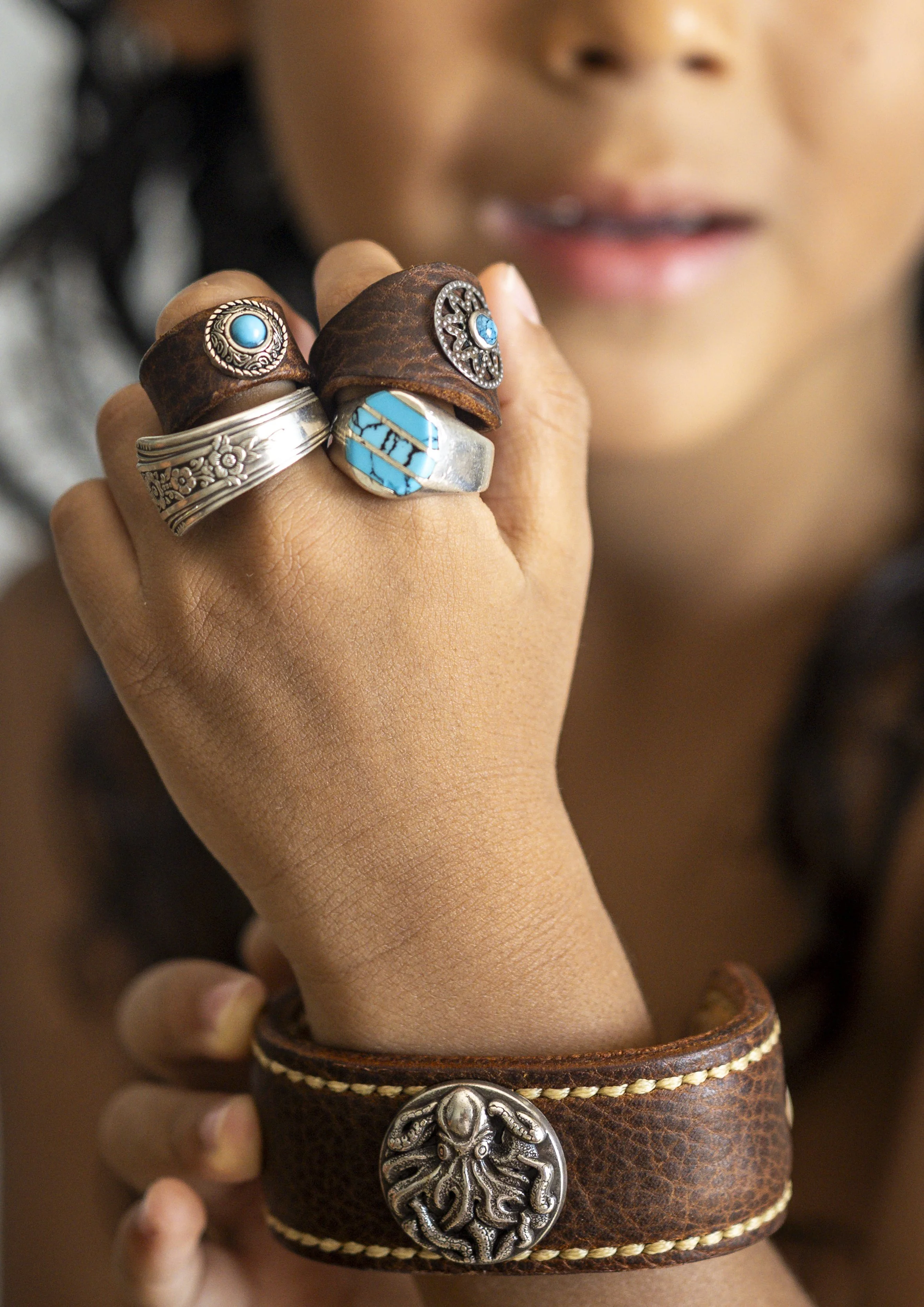 Close-up of a person's hand wearing multiple rings with turquoise and silver accents, and a leather cuff bracelet with a silver octopus emblem. The person's face is blurred in the background.