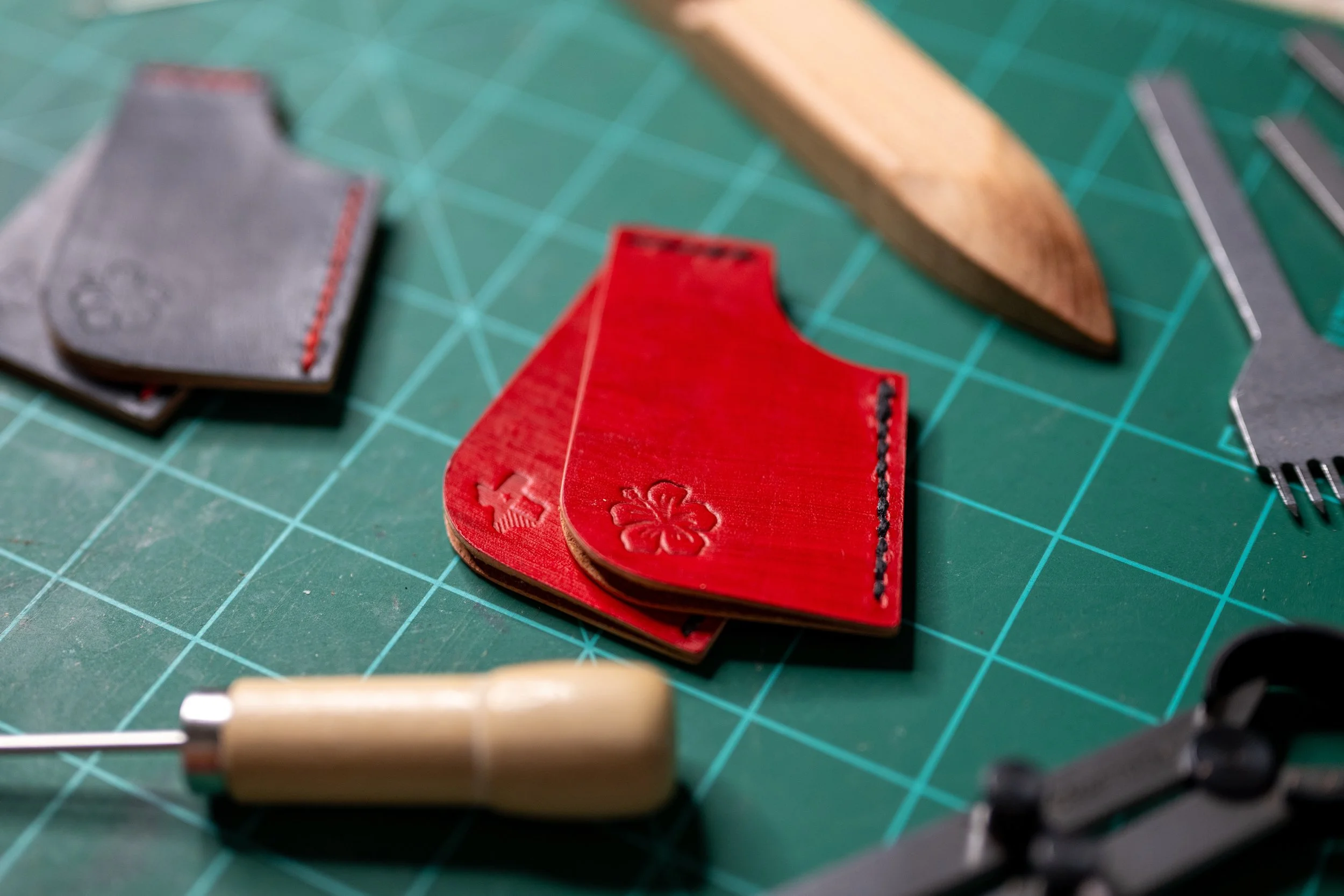 Close-up of leather pieces in gray, red, and tan on a green cutting mat with grid lines, alongside a metal ruler and a rotary cutter handle.