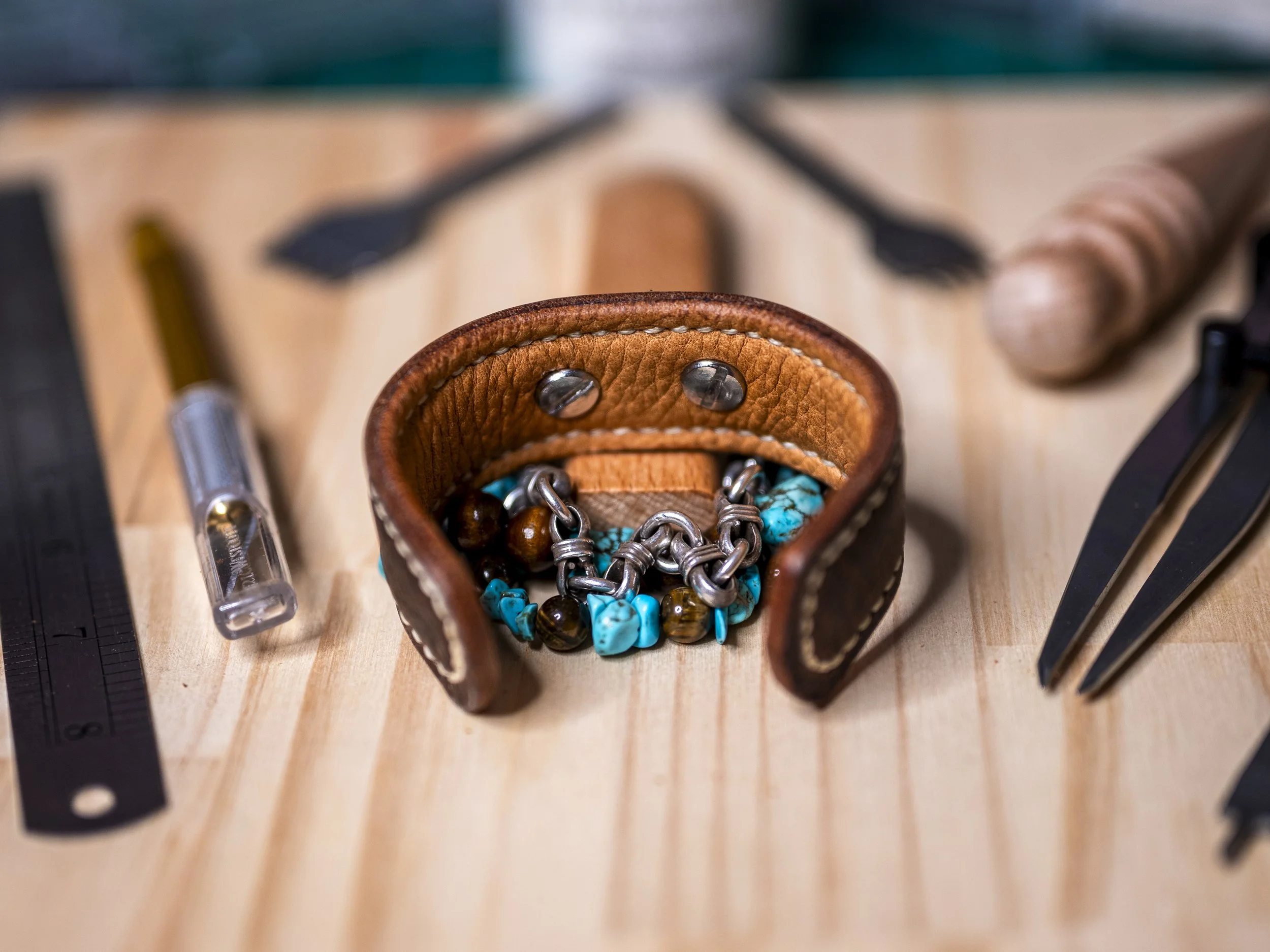 Interior view of the handcrafted Moana brown leather cuff, showing its leather construction and metal hardware, with turquoise and brown beaded jewelry nestled inside and leatherworking tools surrounding it on a wooden workbench.