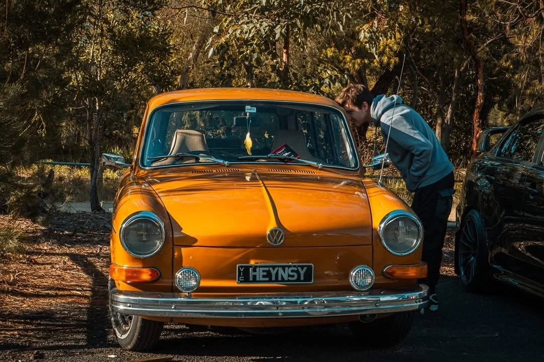 A person looks into the engine of an orange vintage Volkswagen Beetle parked in a wooded area.