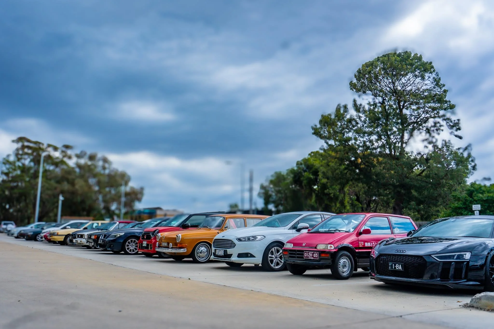 Row of classic and modern cars parked in a lot under a cloudy sky with trees in the background.