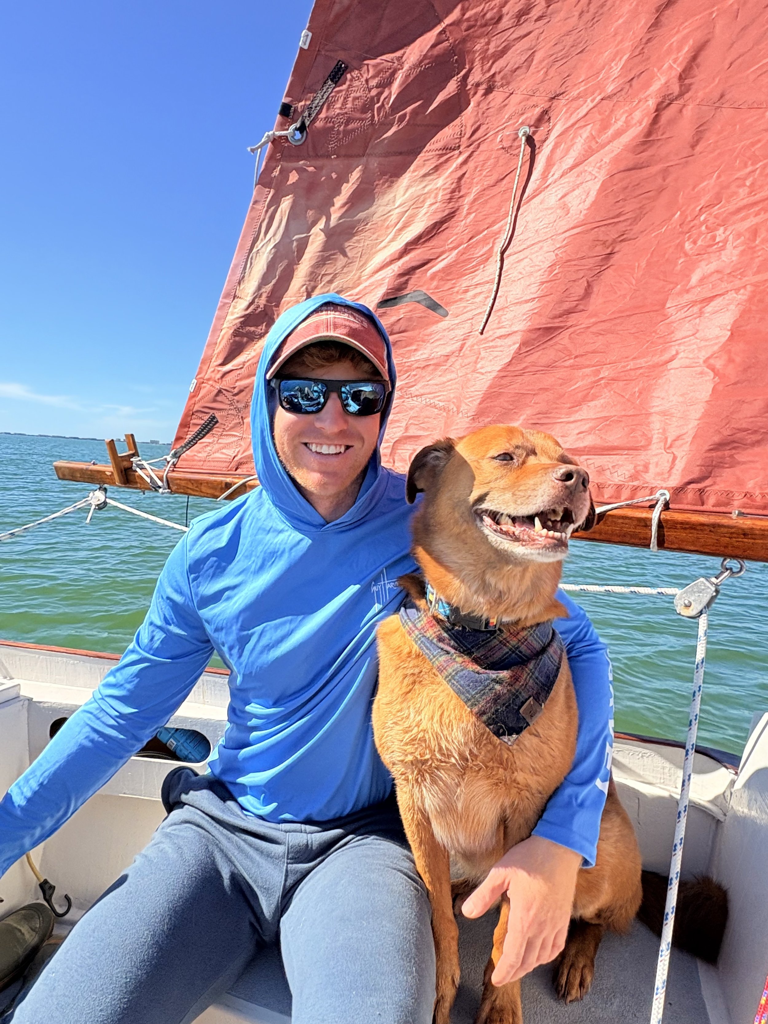 A smiling man wearing sunglasses, a blue hoodie, and gray pants, sitting on a boat. He is holding a happy, brown dog with a plaid bandana around its neck. The boat is on water, with a red sail visible in the background under a clear blue sky.