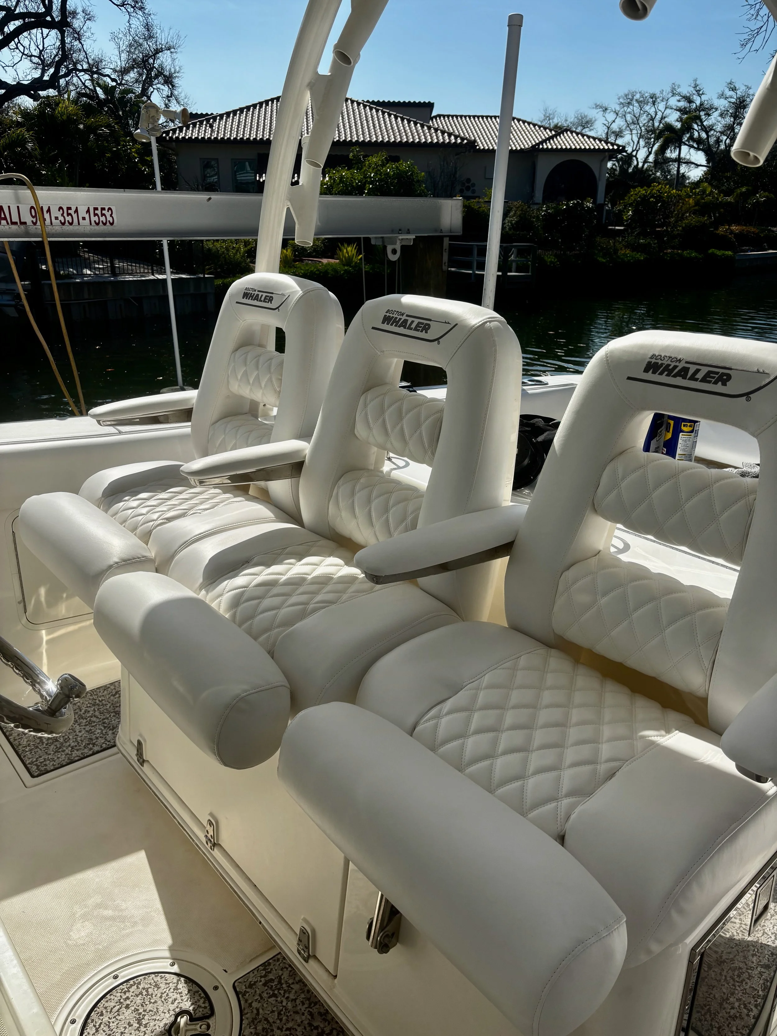 White cushioned boat seats with diamond-pattern stitching, branded 'Boston Whaler', on a boat by a waterway with a house and trees in the background.