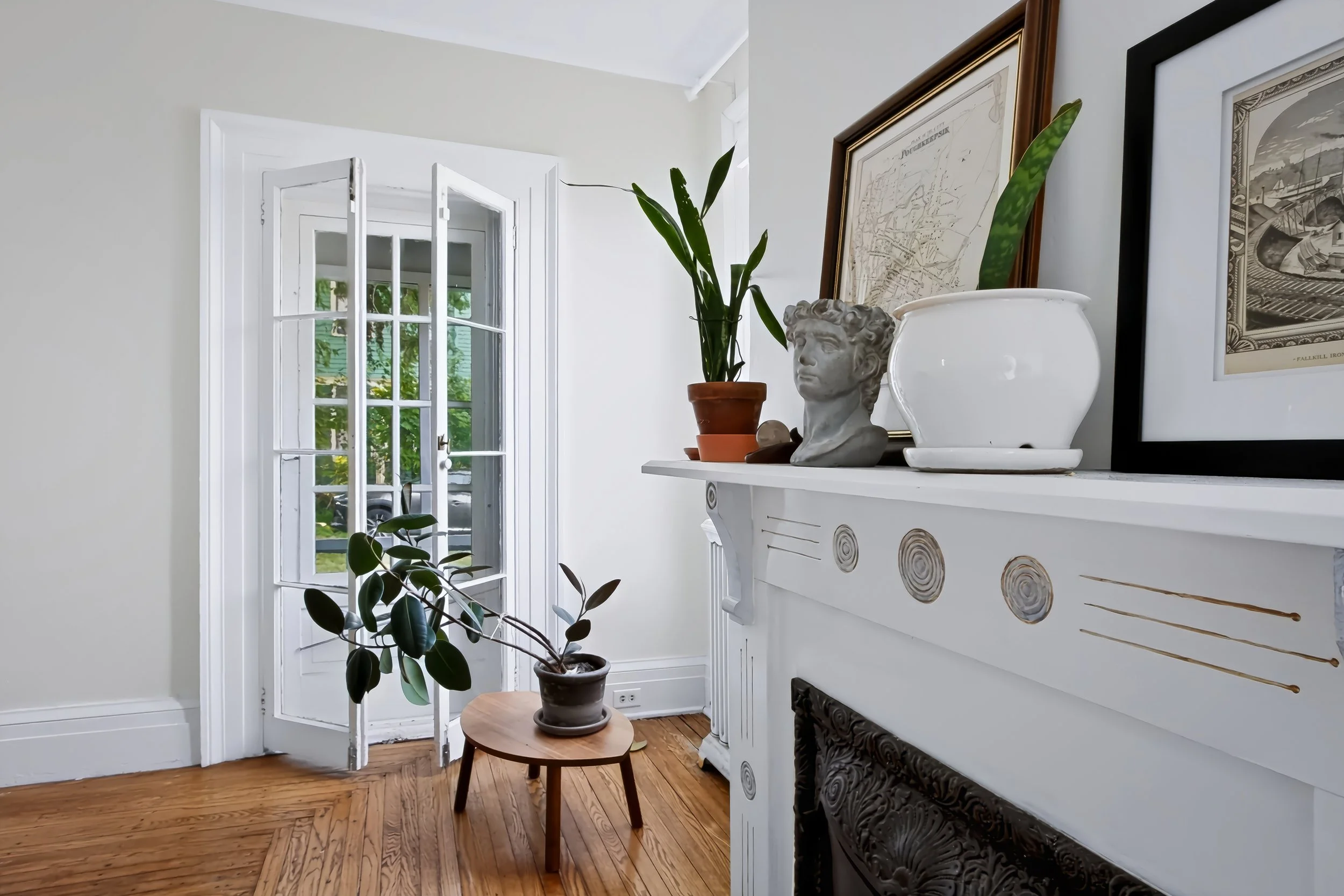 Room with white walls and hardwood floors, decorated with framed artwork, potted plants, and a white fireplace mantel. There are glass-paned doors leading outside, and sunlight is coming into the room.
