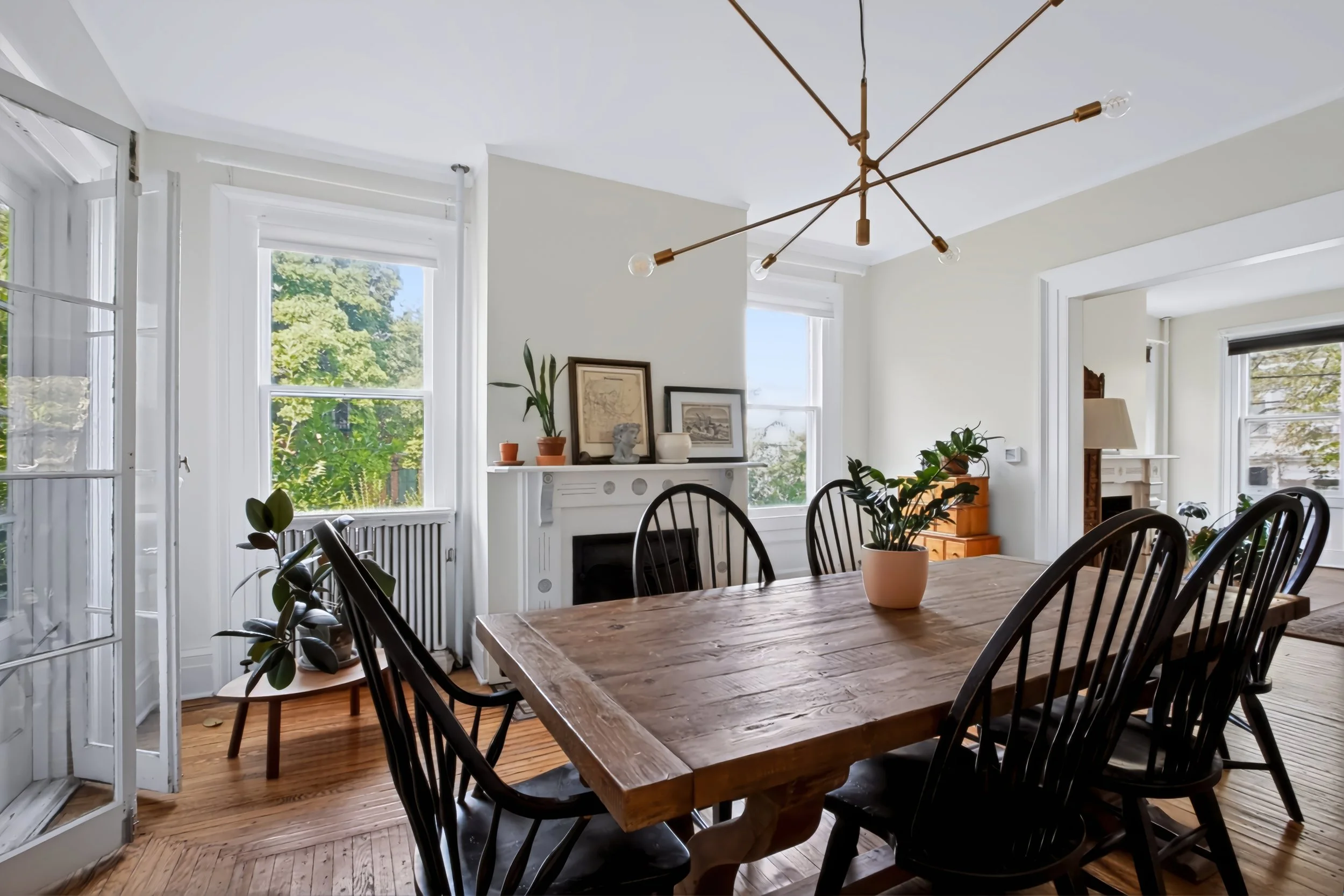 Bright dining room with a wooden table, black chairs, and large windows showing green trees outside. Decor includes framed artwork, potted plants, and a modern gold chandelier.