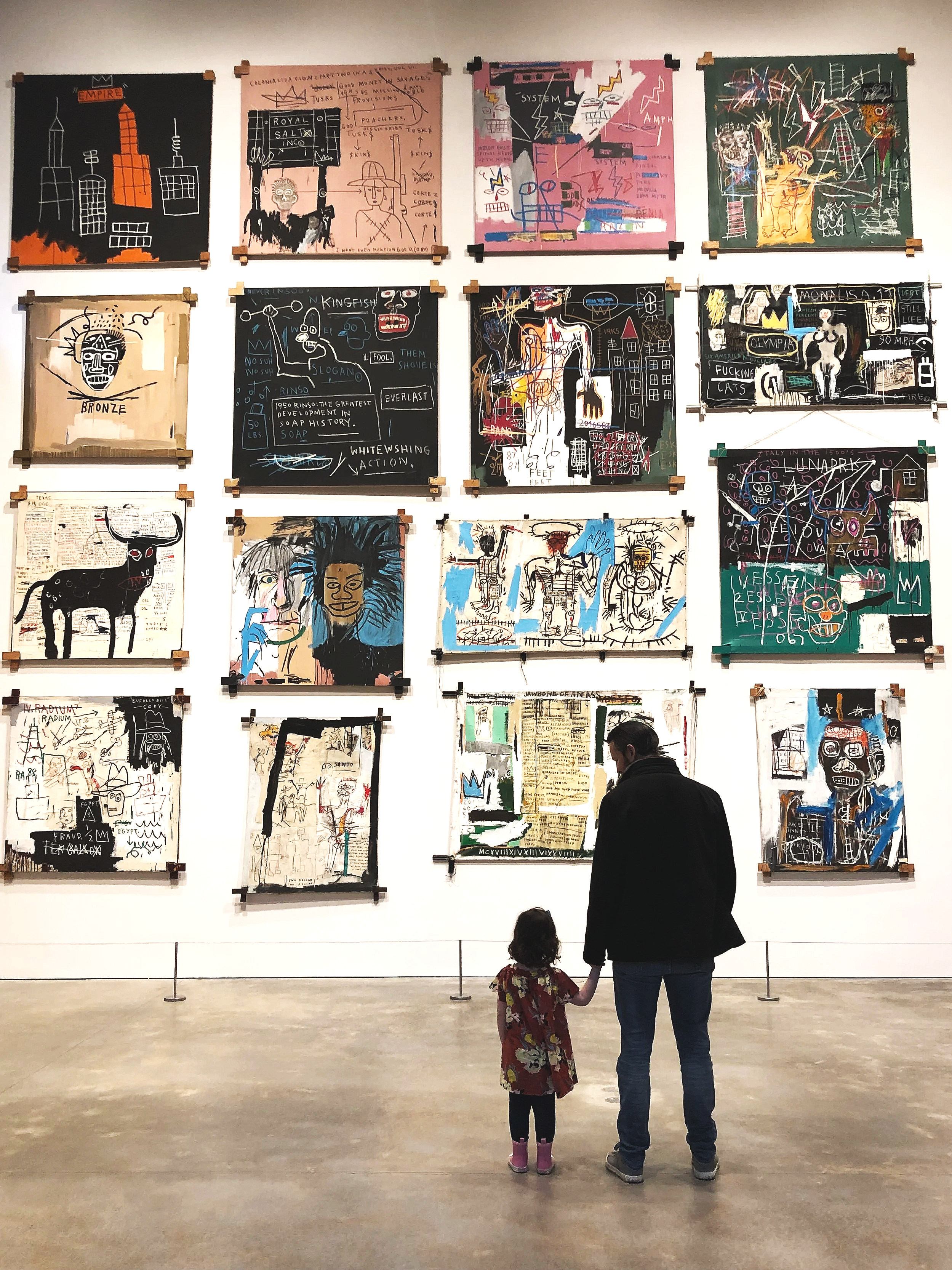 A man and a young girl holding hands stand in front of a large wall of paintings by Jean-Michel Basquiat in the Brant Foundation art gallery. The wall features multiple panels with colorful, chaotic, and expressive designs.