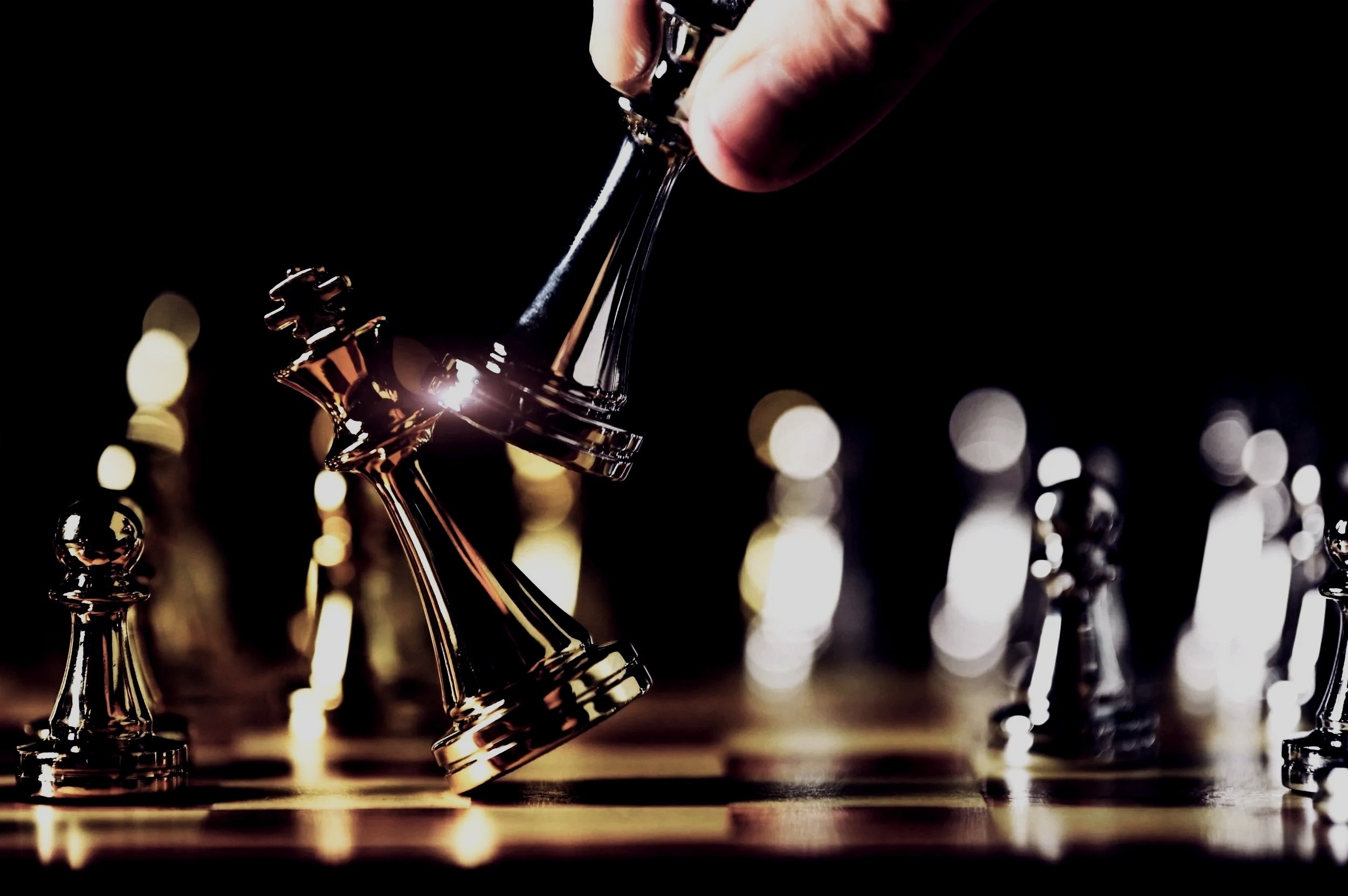 Close-up of a hand moving a shiny metallic king chess piece on a chessboard, with blurred chess pieces and bokeh lights in the background.