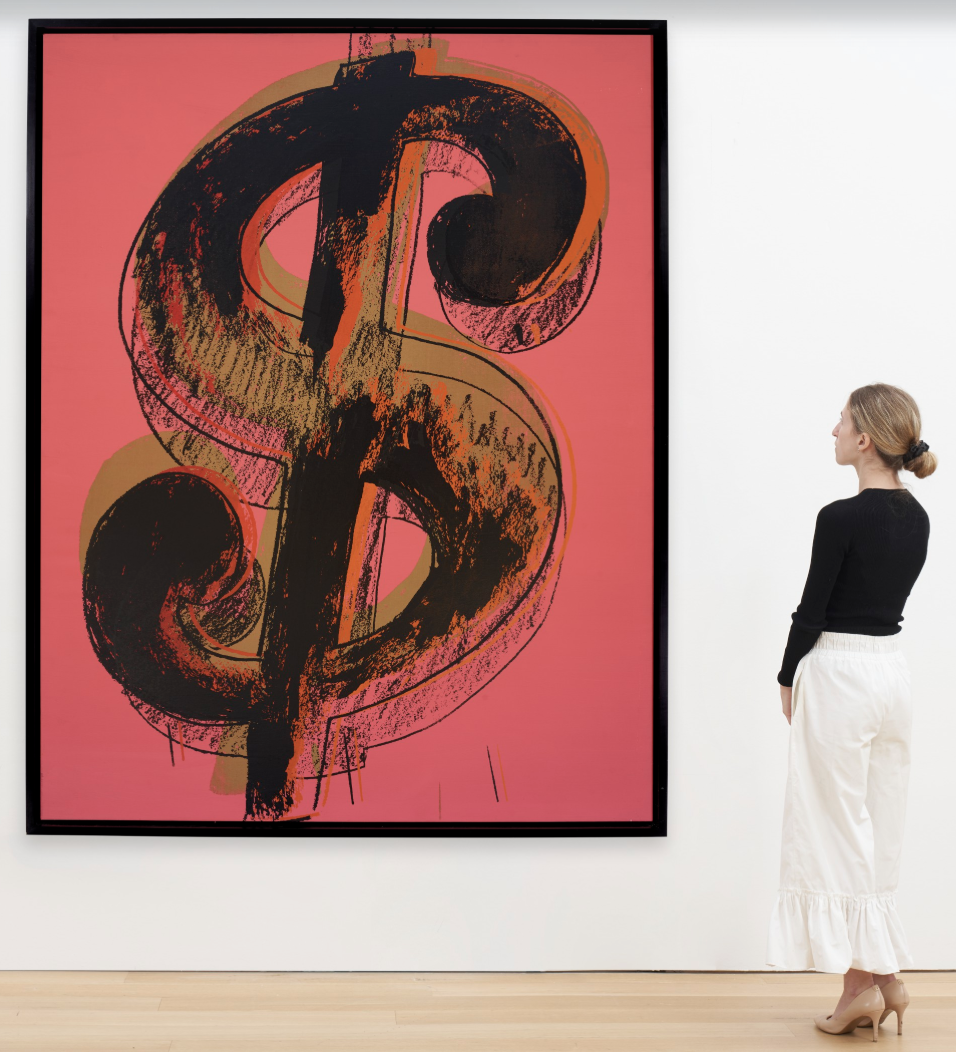 A woman looking at a large red, orange and black dollar sign painting by Pop artist Andy Warhol.