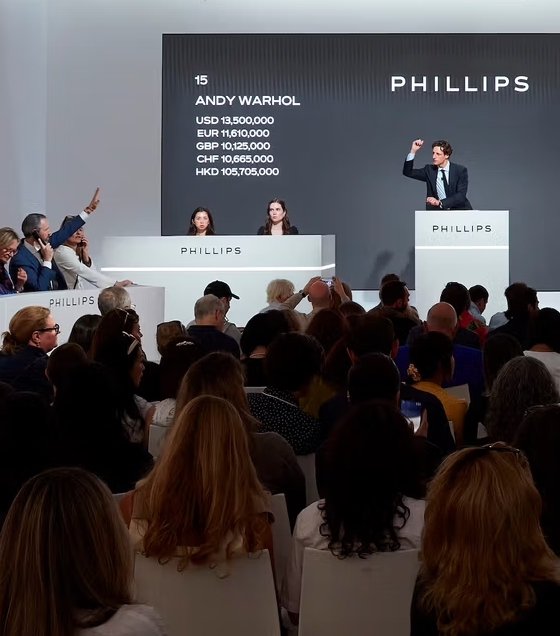 Auction scene with a man standing on a podium and raising his hand, while two women sit at a table behind him. Audience members are seated, some raising their hands, at a Phillips auction for Andy Warhol artwork, with bidding prices displayed on a screen.