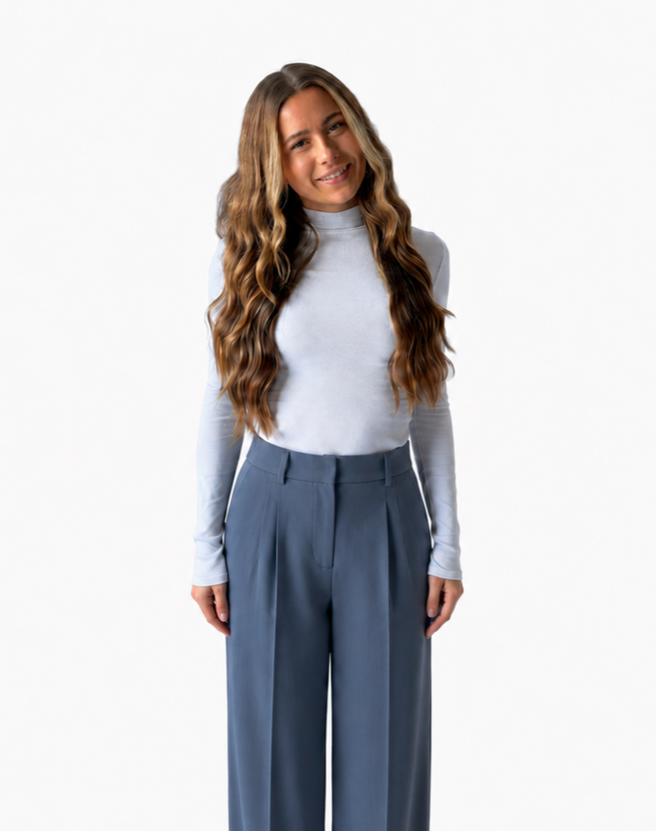 A woman with long wavy hair smiling at the camera, wearing a light gray long-sleeve top and high-waisted blue trousers, standing against a plain white background.