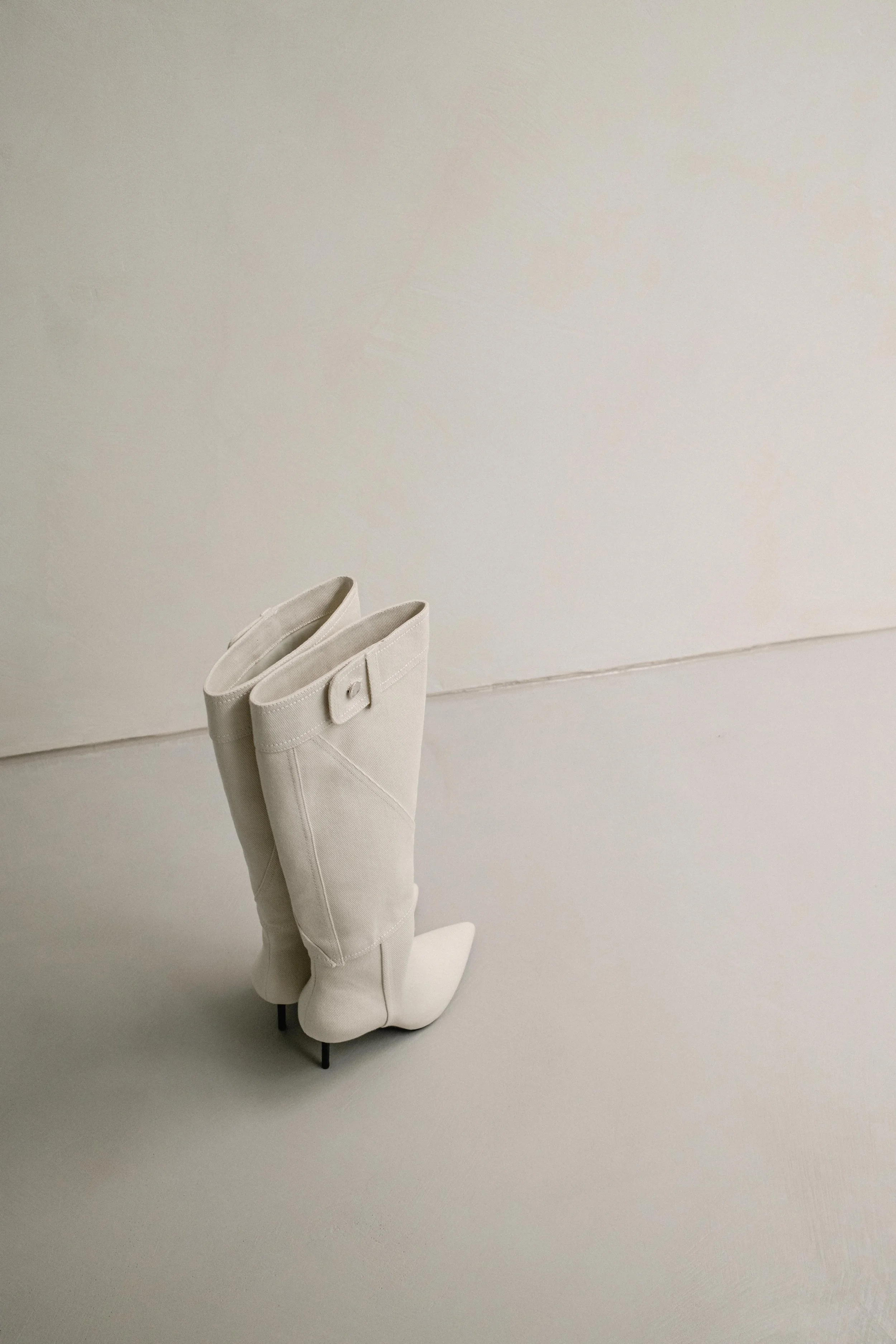 A pair of white high-heeled boots made of a soft, textured material, standing upright on a small black stand with thin legs against a plain light-colored background.