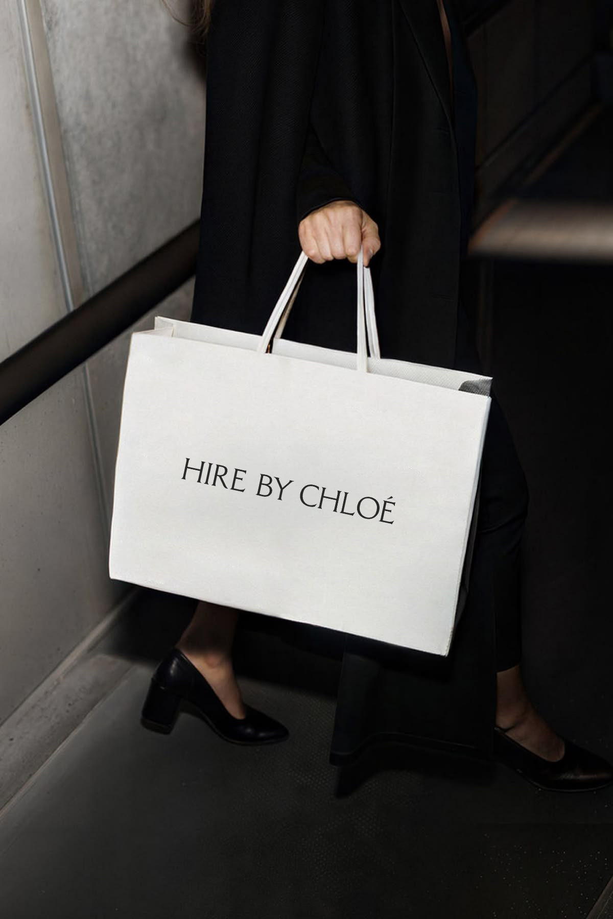 A person holding a white shopping bag with black text that reads 'HIRE BY CHLOÉ,' wearing black high-heeled shoes, black pants, and a black blazer in an elevator.