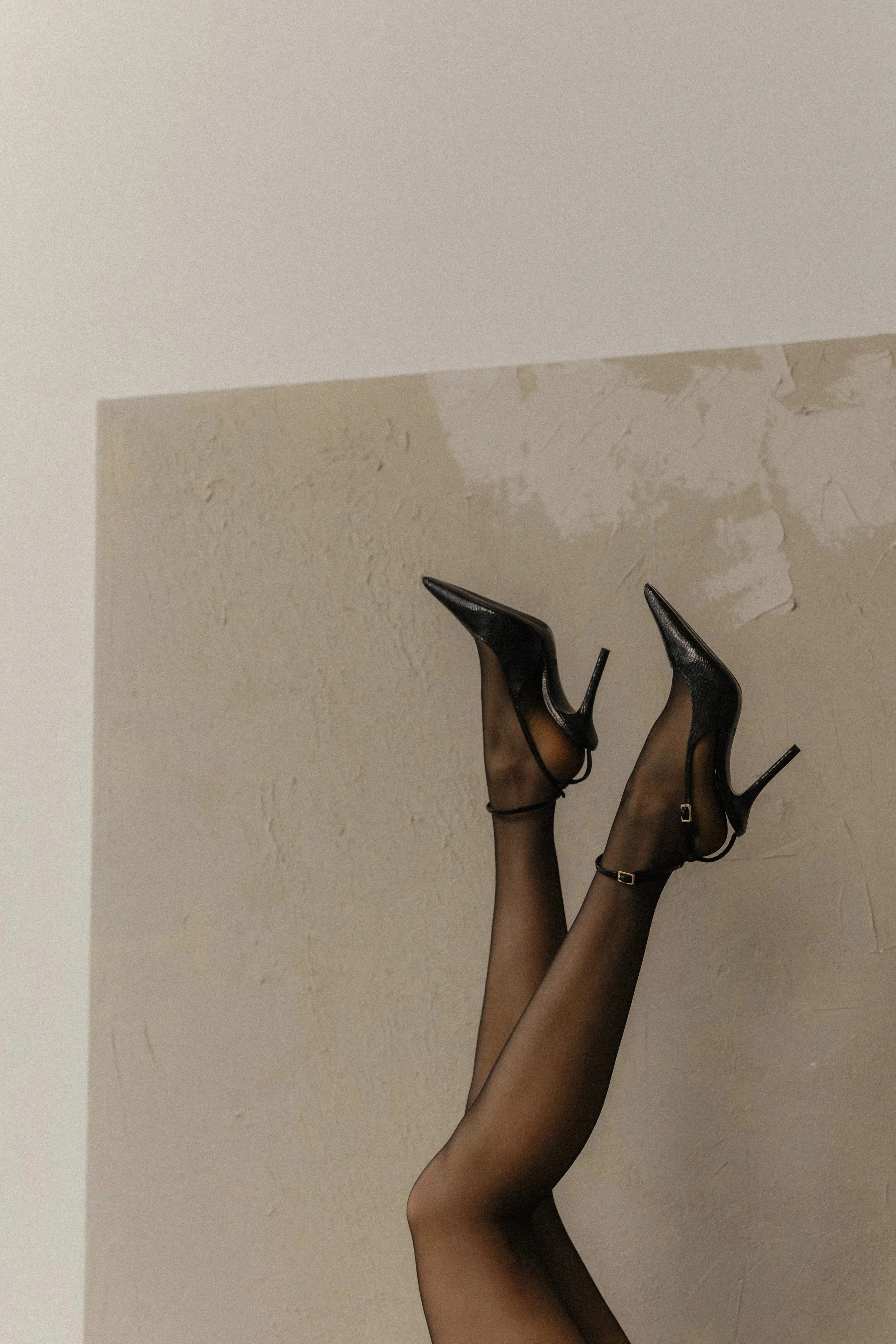Close-up of a woman’s legs in black high heels and sheer stockings, upside down with ankles against a light-colored wall.