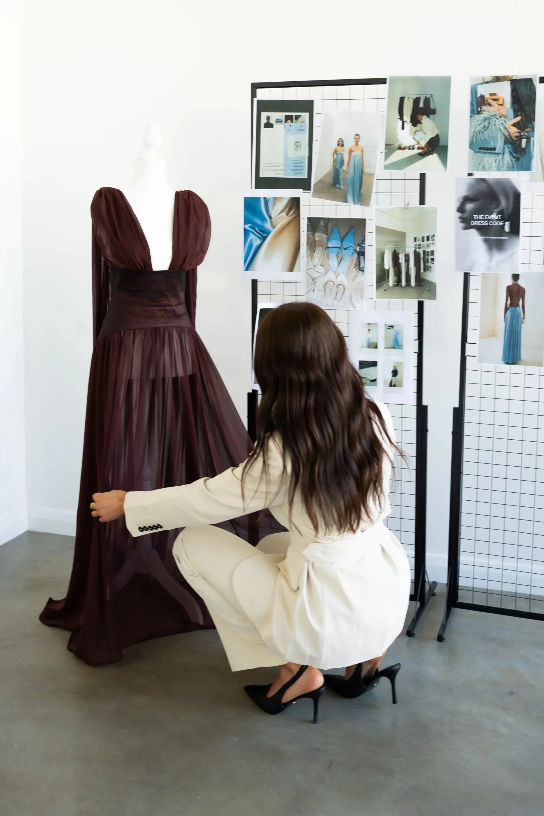 A woman in a white suit with long, wavy hair kneels on the floor adjusting a maroon evening gown on a mannequin. The gown has puffed sleeves and a pleated bodice, with a sheer, flowing skirt. Behind her is a display board with fashion photographs and sketches, and another woman with long hair is partially visible, crouching in front of the display.