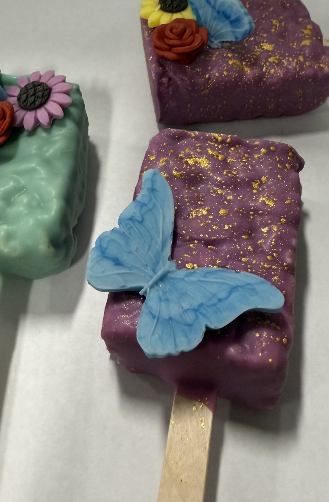 Close-up of a purple ice cream bar with gold flecks and a blue butterfly-shaped decoration on top, on a wooden stick. Partially visible is another ice cream bar with light green coating and flower decorations.