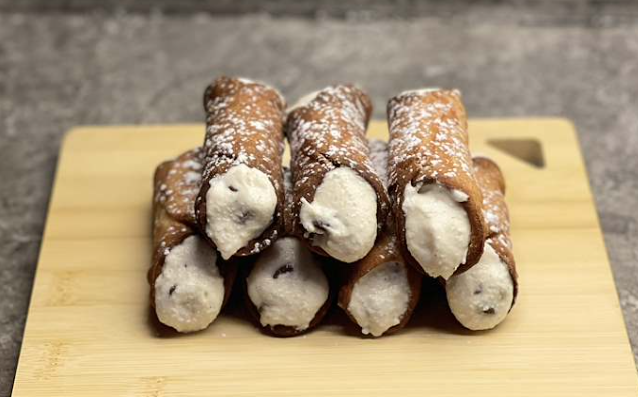 Six ice cream-filled cannoli with powdered sugar on top, arranged on a wooden cutting board.