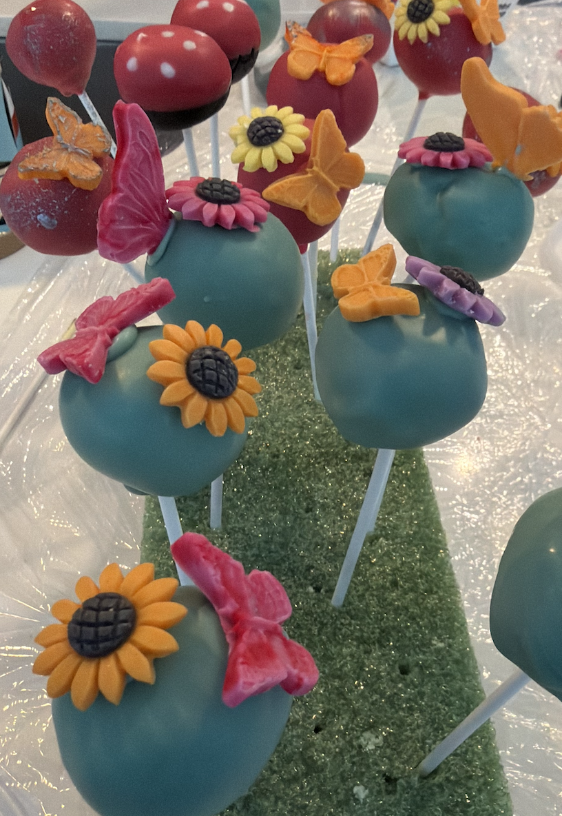 Cake pops decorated with colorful flowers, butterflies, and bows, arranged on a tray with a green textured strip, resembling a garden scene.