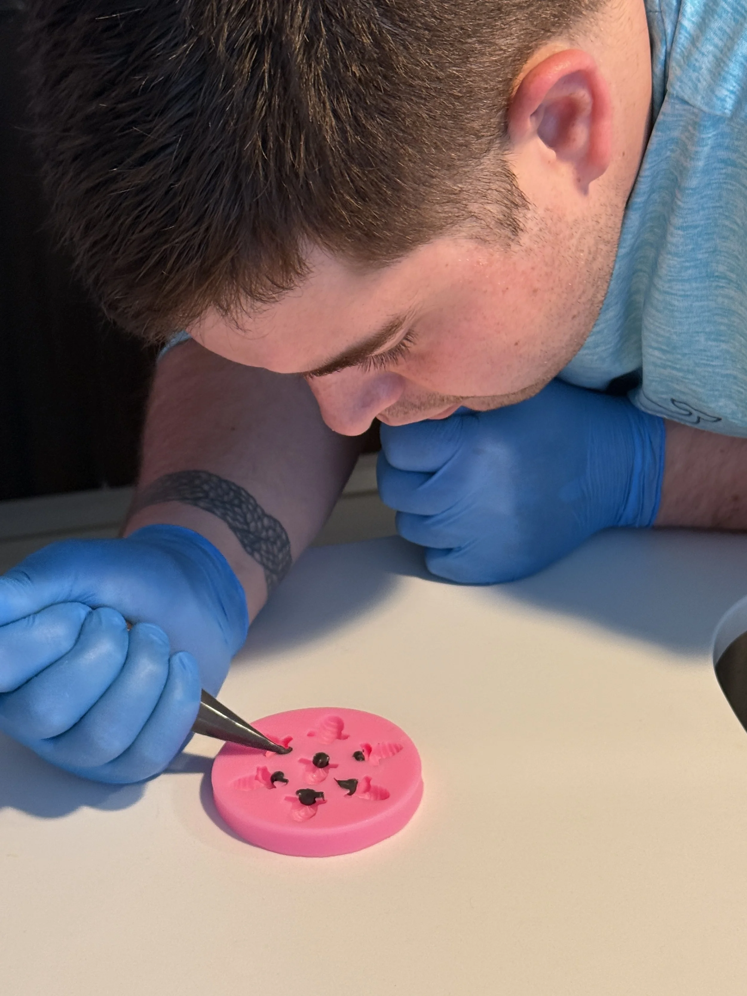 A person with short dark hair and wearing blue gloves is using a tool to place small black objects into holes of a pink silicone mold on a white surface.