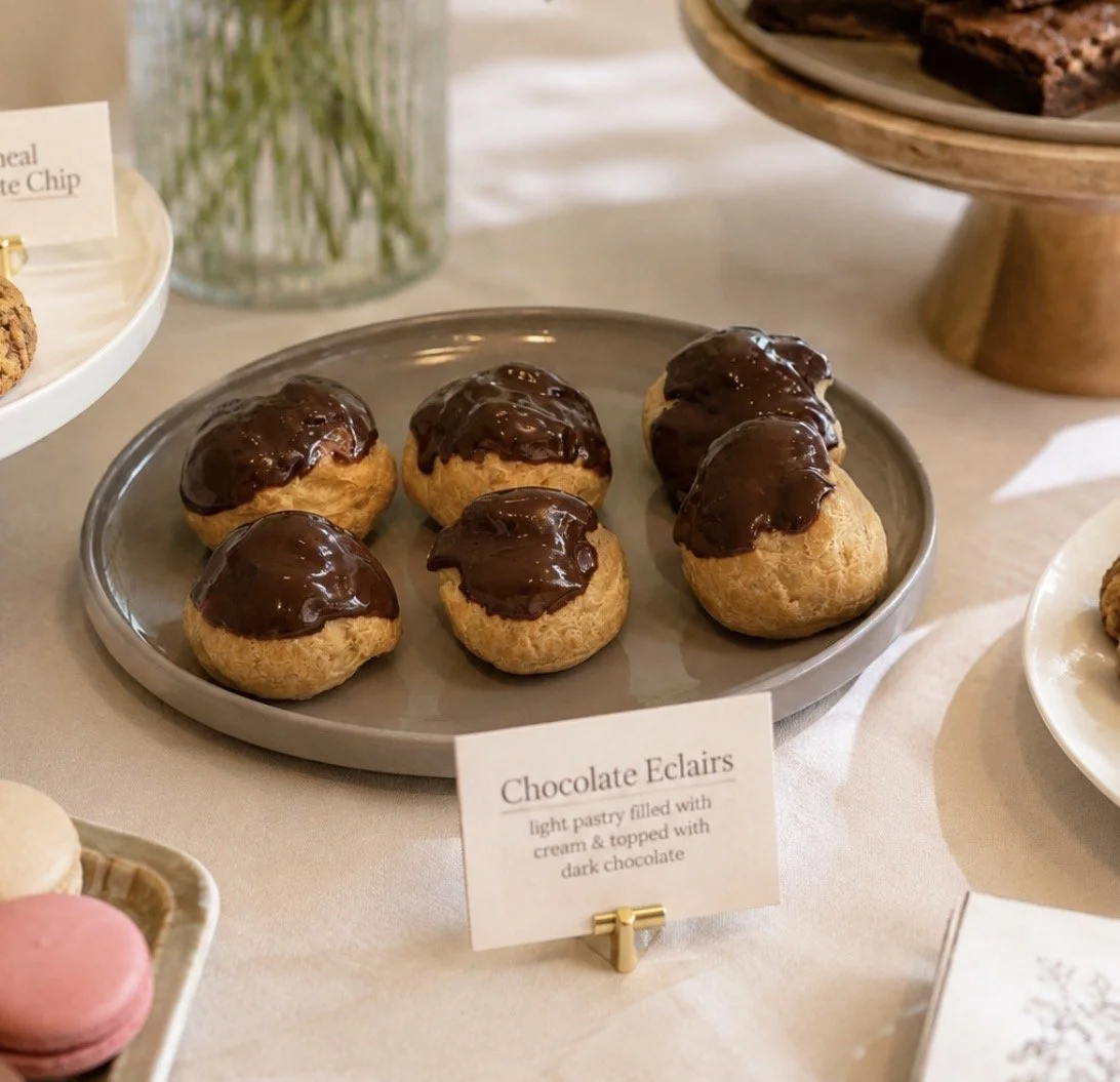 Plate of chocolate éclairs with light pastry filled with cream and topped with dark chocolate, on a table with other desserts and a glass vase in the background.