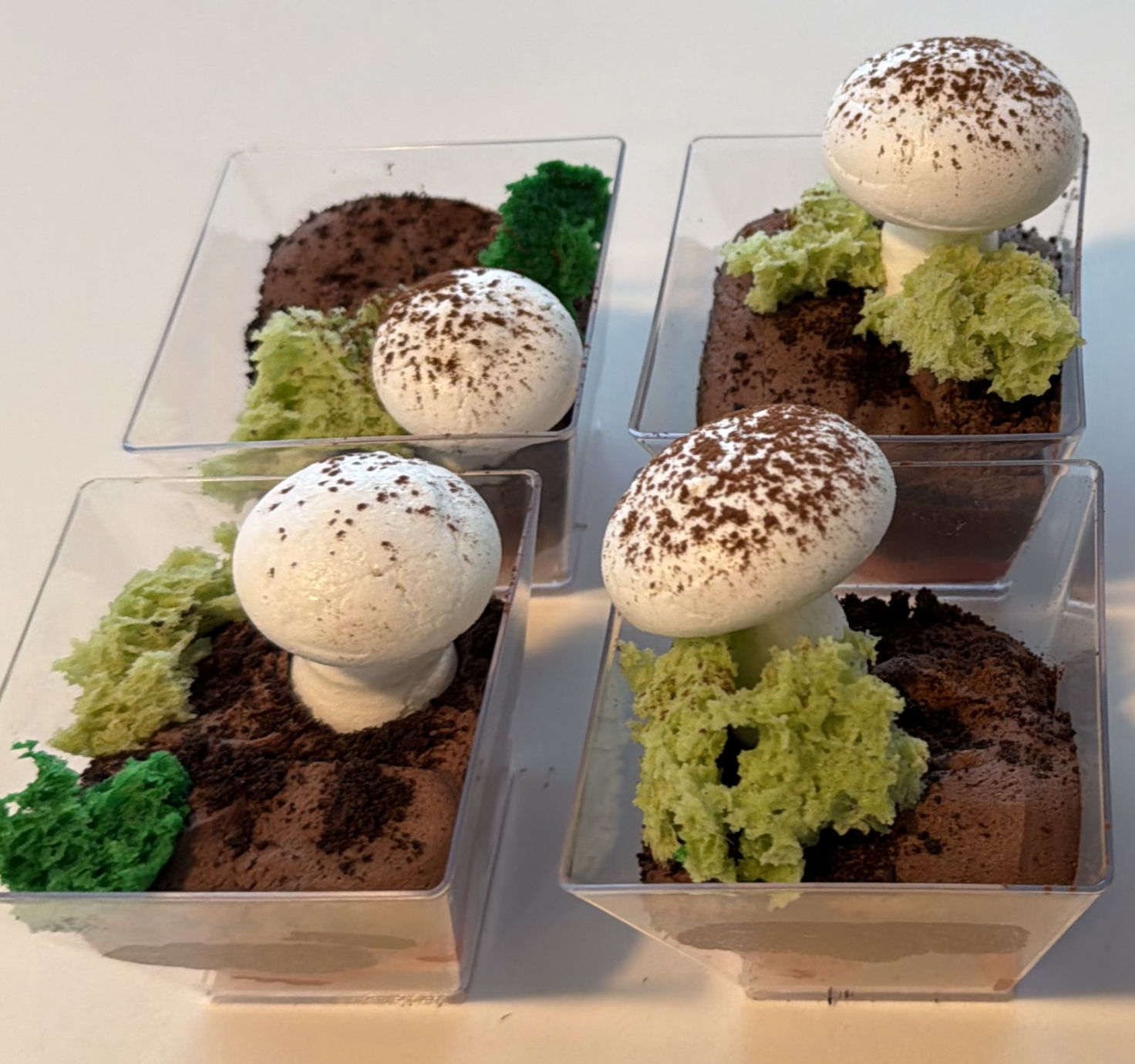Four clear square dessert cups filled with chocolate mousse, decorated with whipped cream, green sponge cake, and chocolate sprinkles.