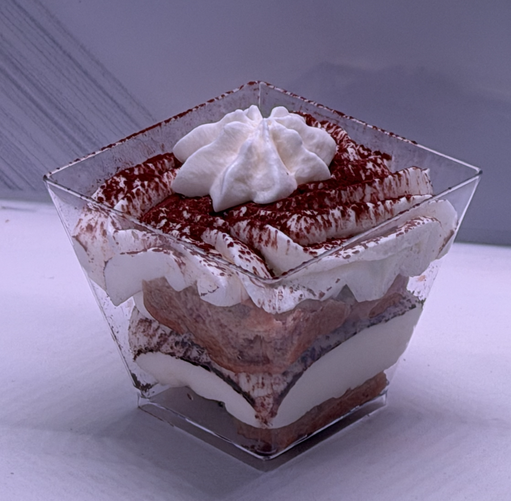 A clear square dessert cup containing layered tiramisu with whipped cream and cocoa powder on top.