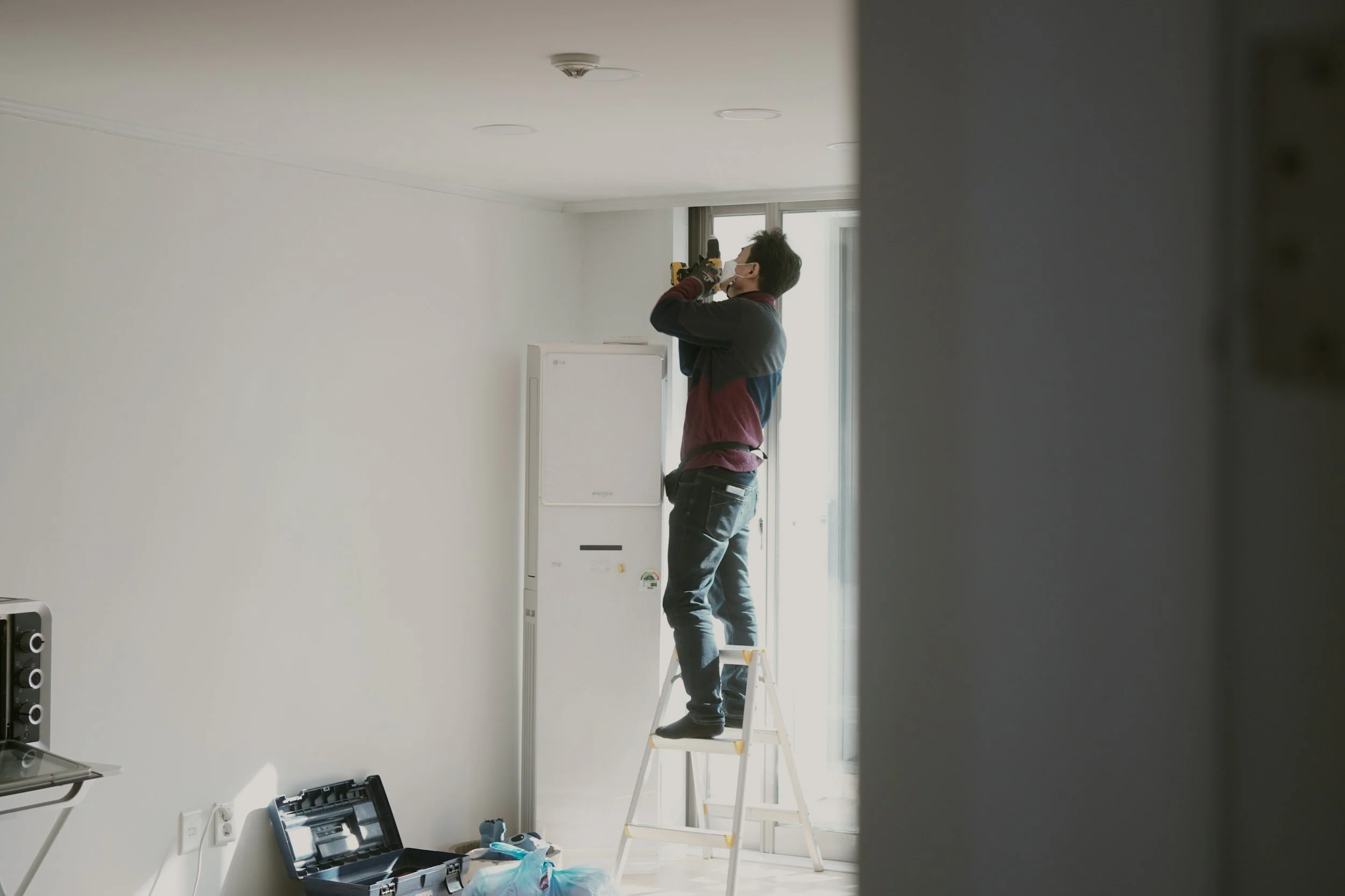 A person standing on a step ladder installing or repairing something on the ceiling in a room with white walls and a window. The individual is wearing a face mask and gloves, holding a power drill.